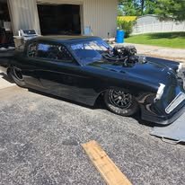 Black vintage car parked in a driveway with a supercharged engine and custom modifications.