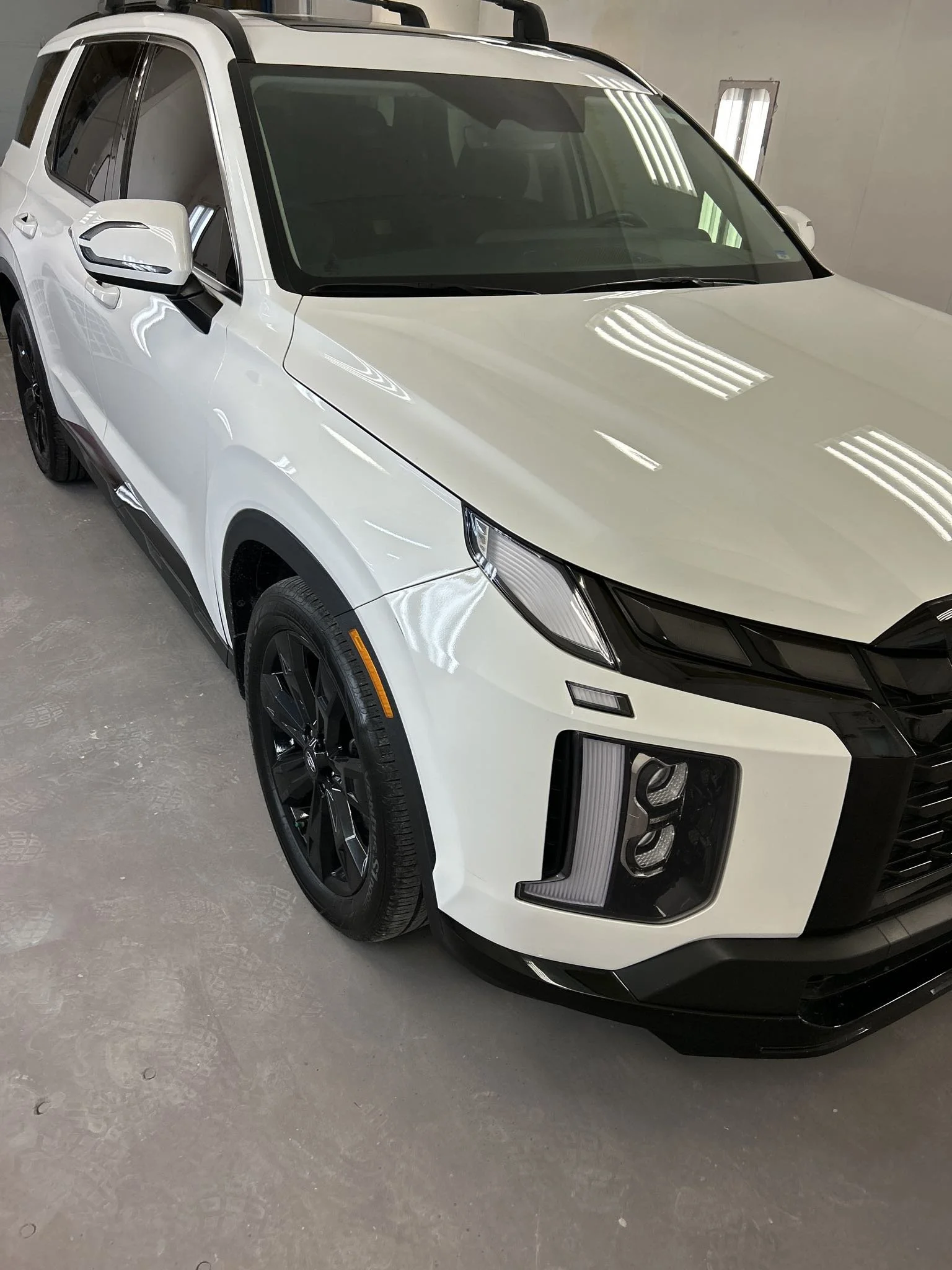 White SUV parked indoors with black wheels and a black front grille, reflecting ceiling lights on the hood.