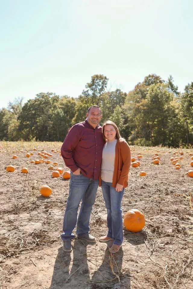 A man and woman standing in a pumpkin patch with pumpkins scattered around on the ground, trees in the background, and clear sky above.