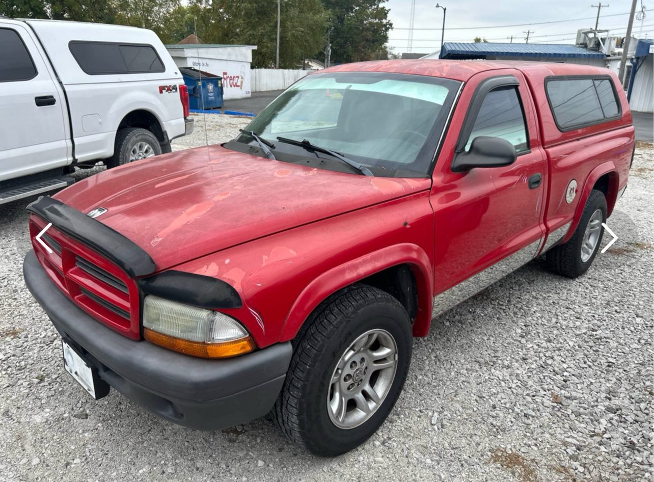 A red pickup truck with a canopy parked on a gravel lot next to a white vehicle. The truck has black fender flares and an older appearance with some paint wear.