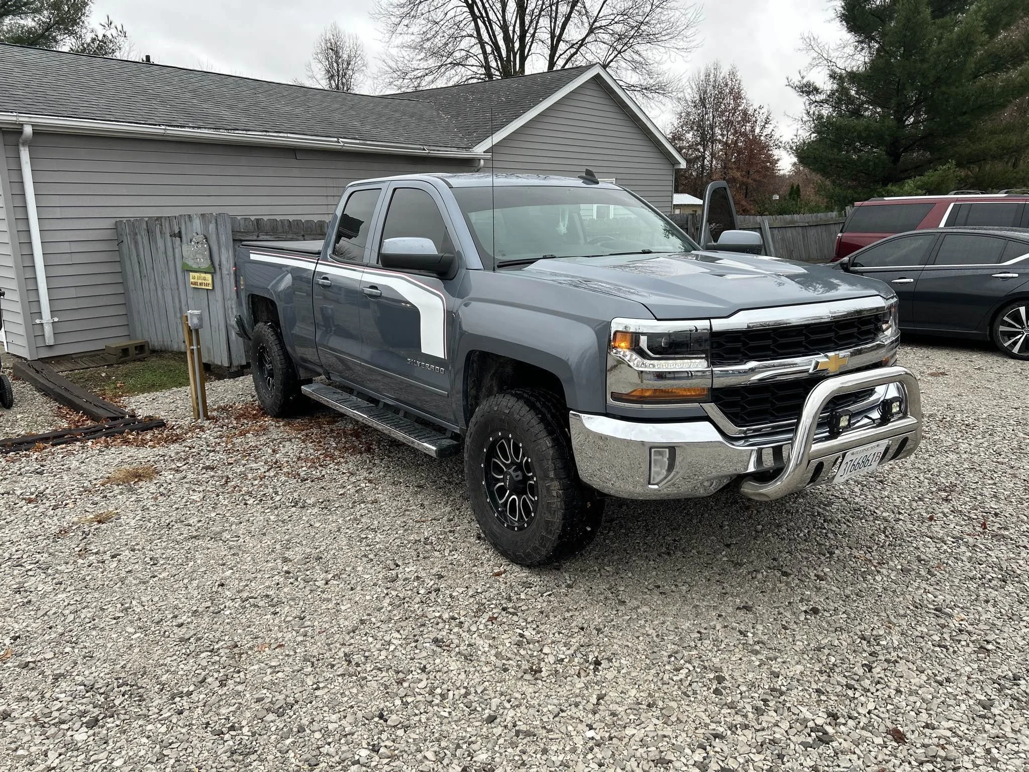 A gray Chevrolet Silverado pickup truck with black wheels parked in a gravel lot next to a gray house, with other vehicles and trees in the background.