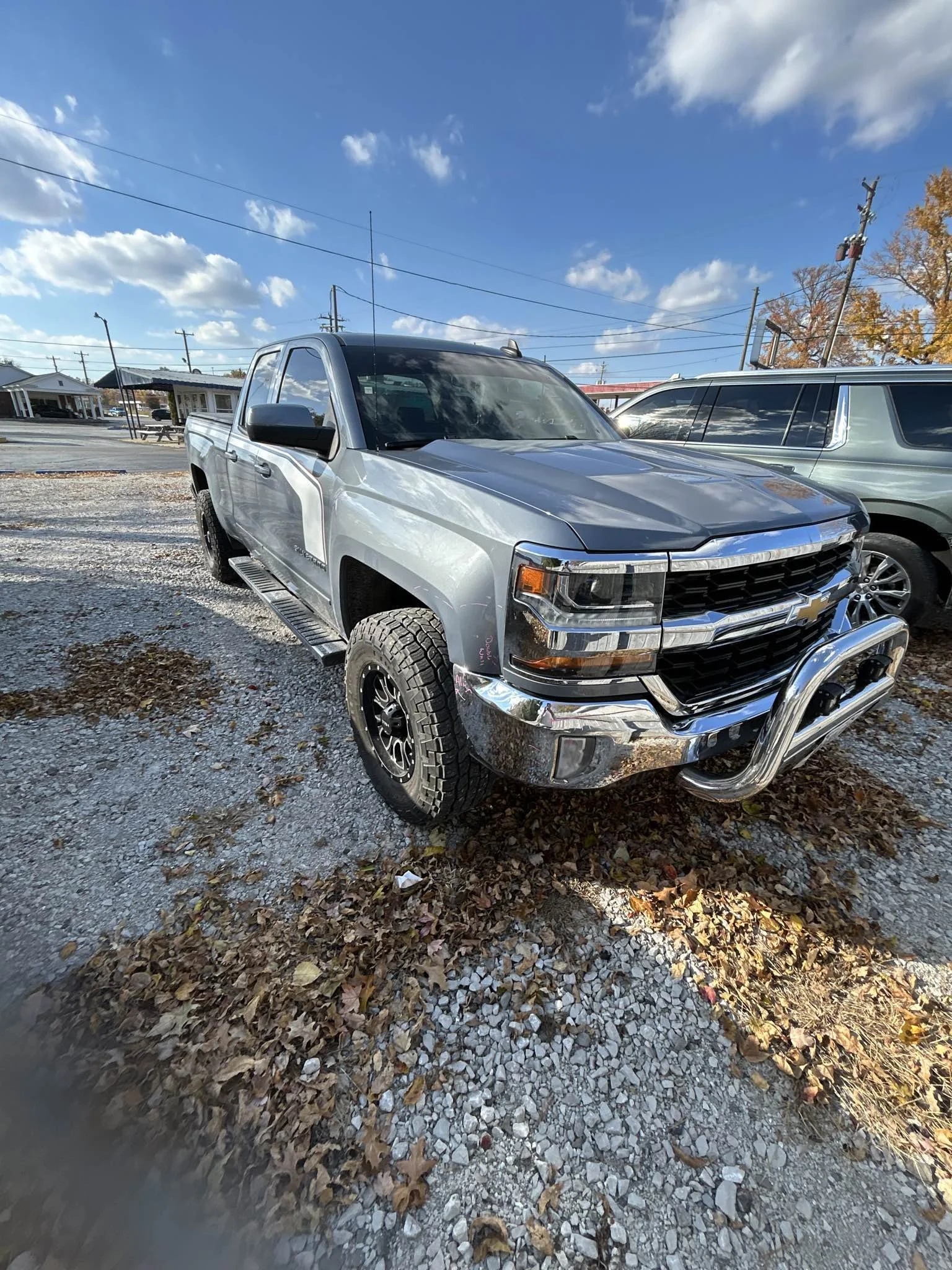 Silver Chevrolet Silverado pickup truck parked on a gravel lot with leaves, under a partly cloudy blue sky.