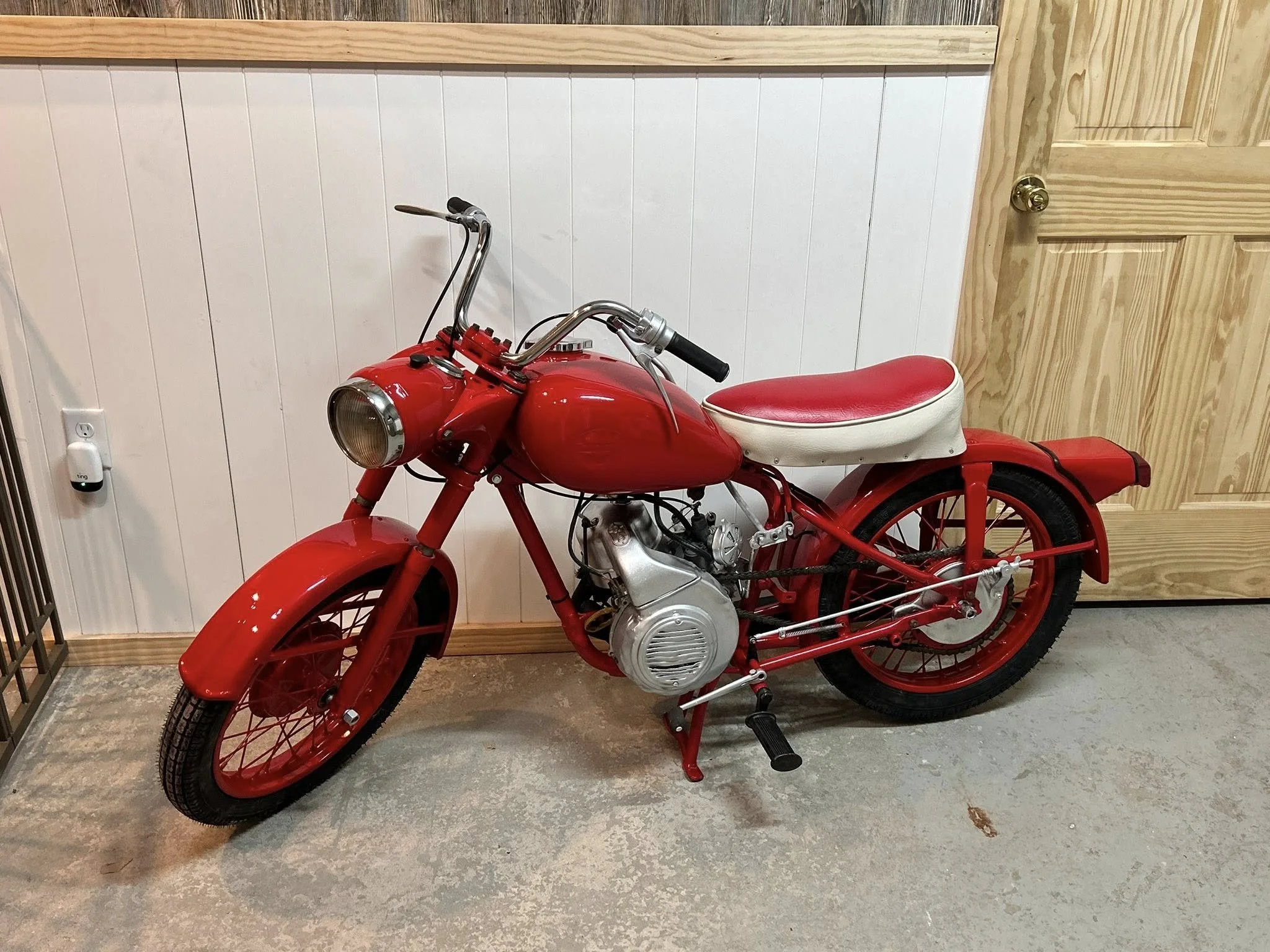 A vintage red motorbike with a white seat parked indoors against a wall with white paneling and a wooden door.