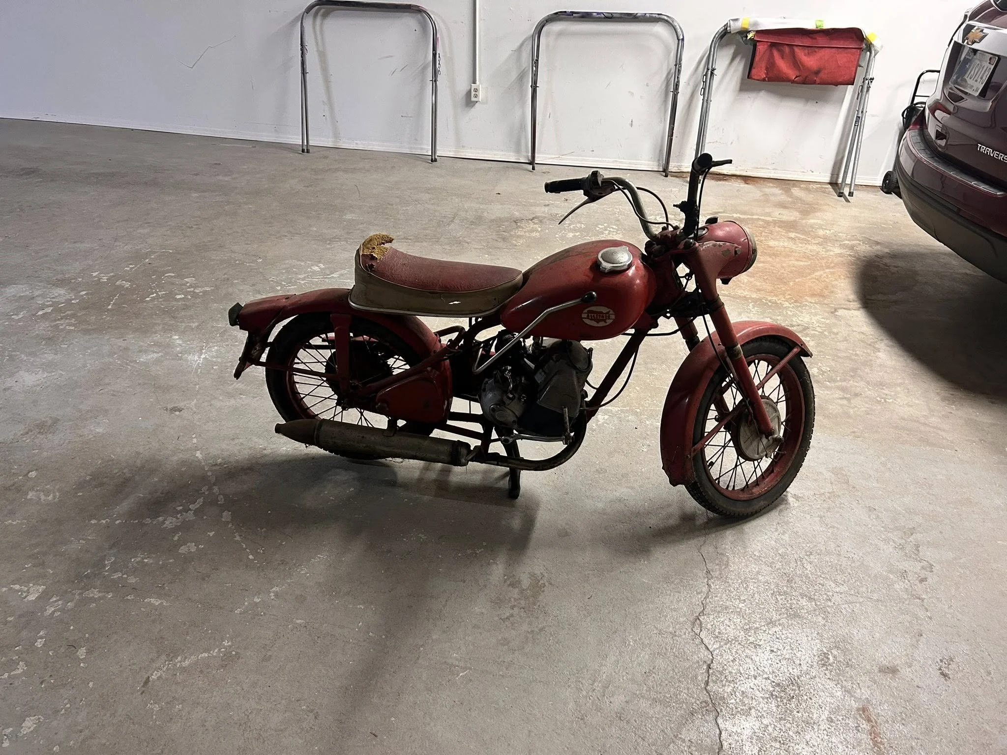 An old, red vintage motorcycle parked in a garage with a damaged seat and visible wear, next to a car.