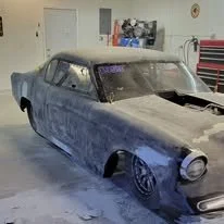 Dusty, old black car in a garage with tools and posters on the wall.