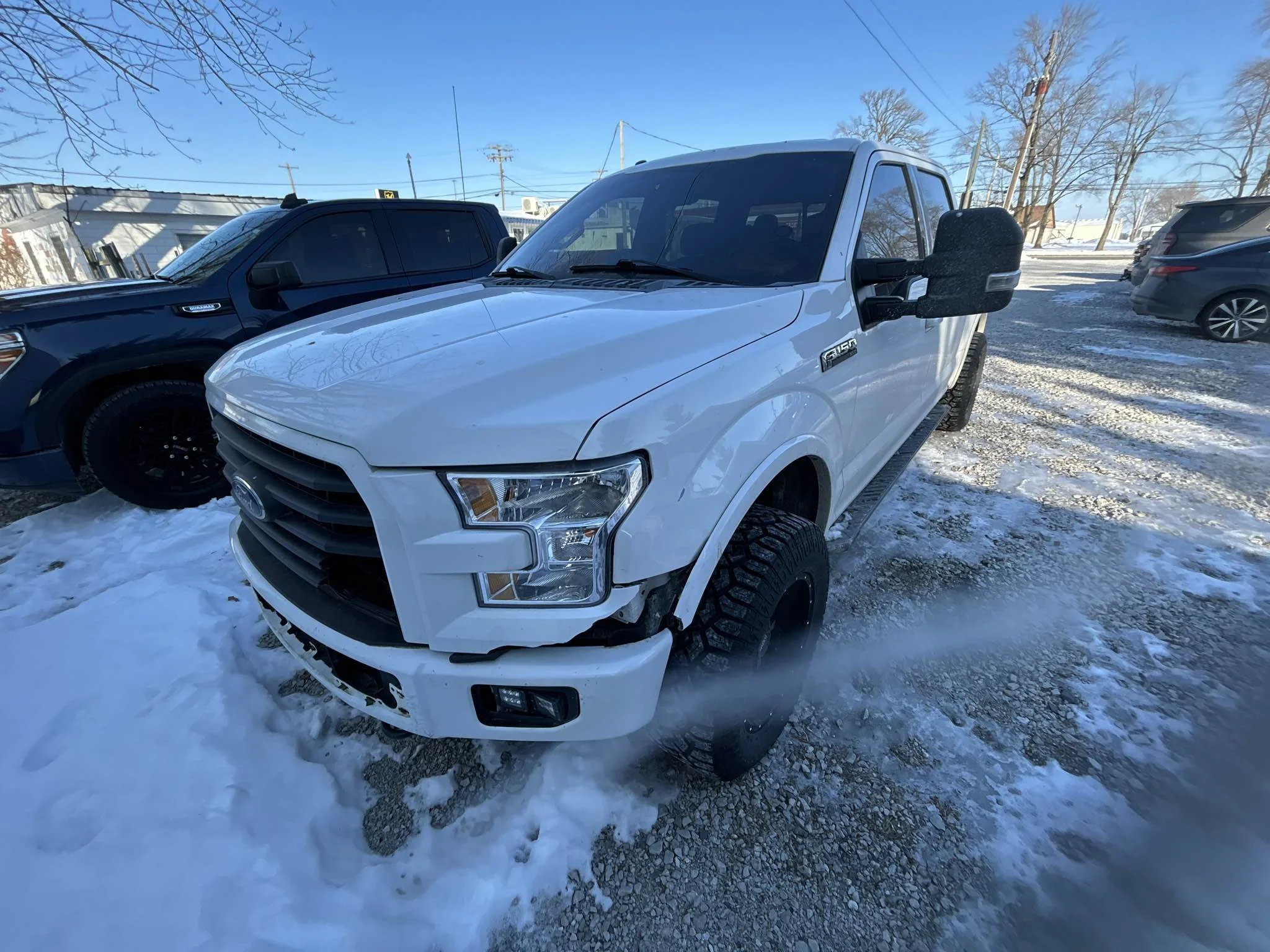 White Ford F-150 pickup truck parked on snowy ground, with damage to the front bumper and tire area, in a lot with other vehicles and a clear blue sky.
