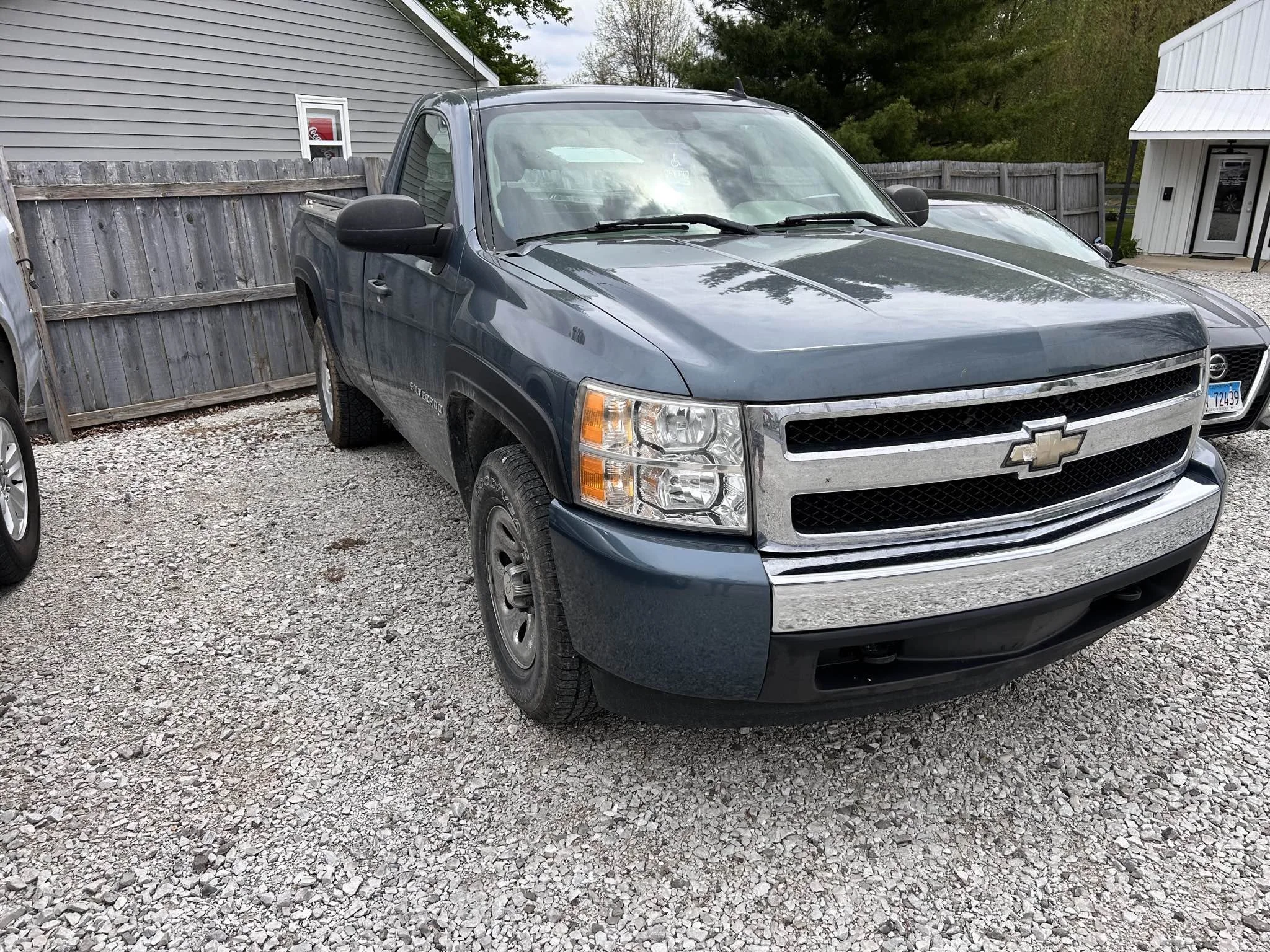 Blue Chevrolet Silverado pickup truck parked on gravel lot, with wooden fence and other vehicles nearby, and a residential building in the background.