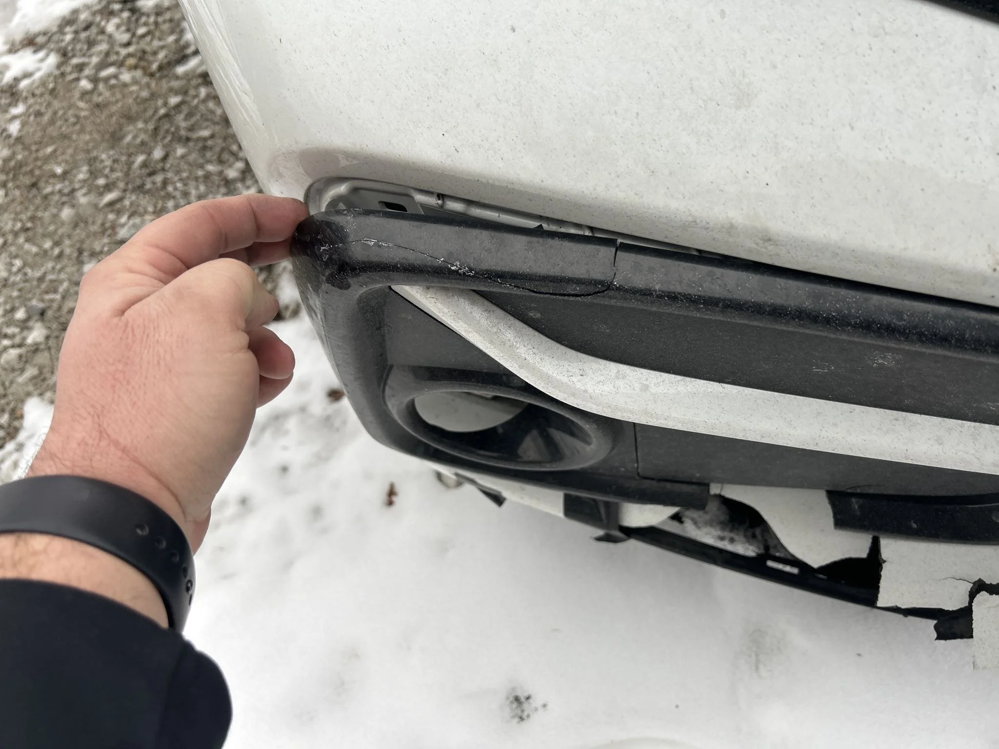 Person inspecting the rear bumper of a white vehicle, focusing on a scuffed black plastic corner near the taillight, with snow on the ground.