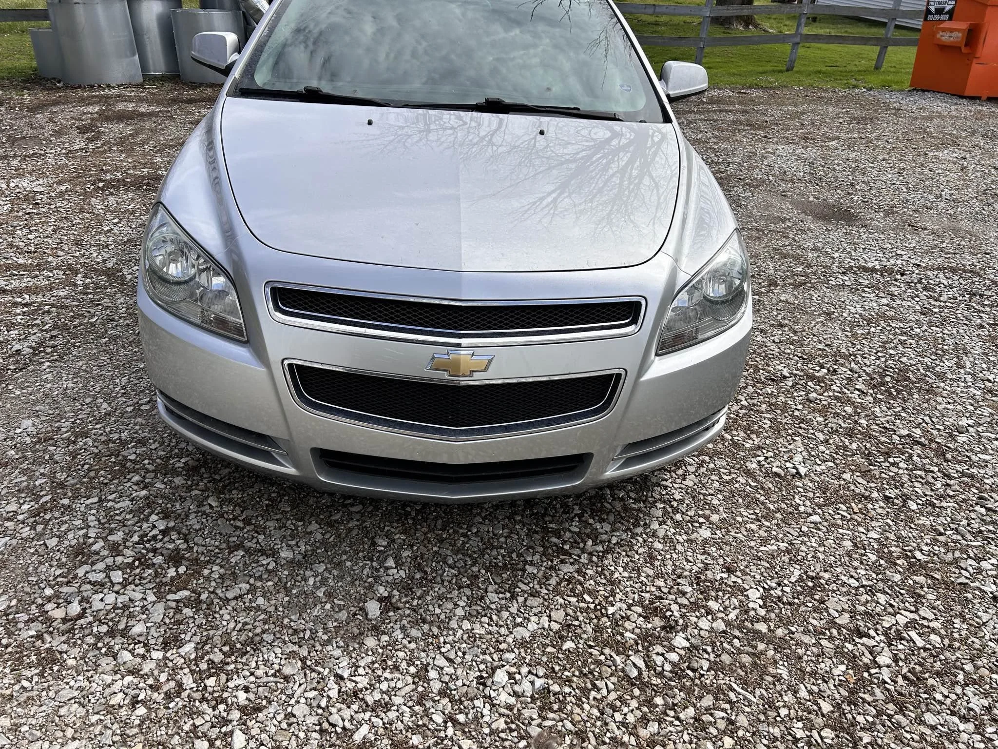 Front view of a silver Chevrolet sedan parked on a gravel surface.