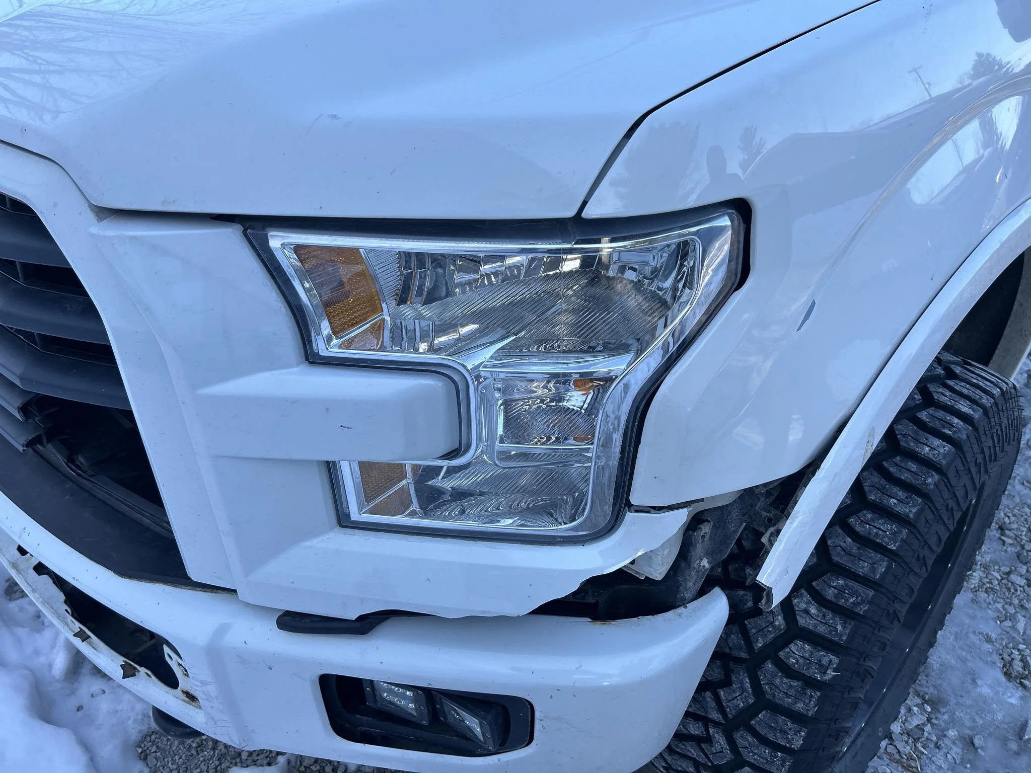 Close-up of the front left corner of a white pickup truck with visible damage near the headlight and bumper, parked on a snowy surface.