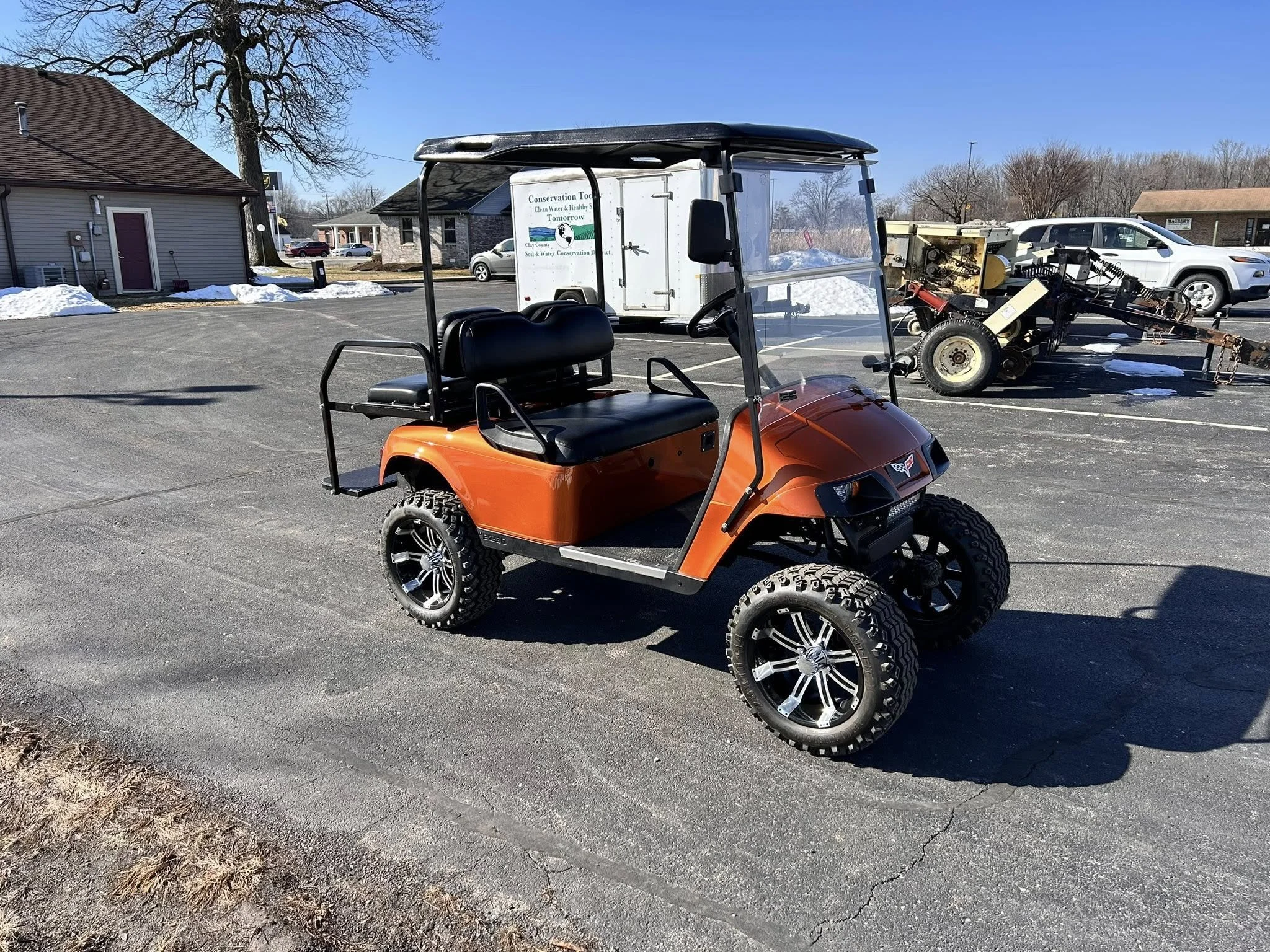 An orange golf cart with black seats and large tires parked on asphalt in a parking lot with some snow and buildings in the background.