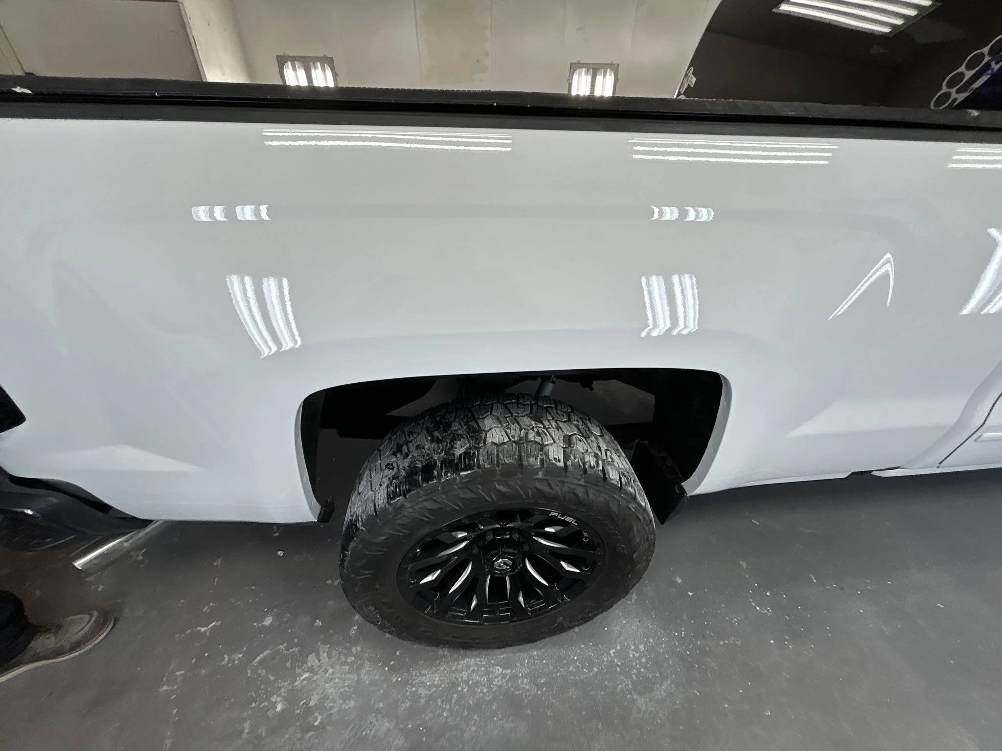 Close-up view of the side of a white pickup truck, showing a black off-road tire with black rim and the vehicle's wheel well, inside a well-lit garage.