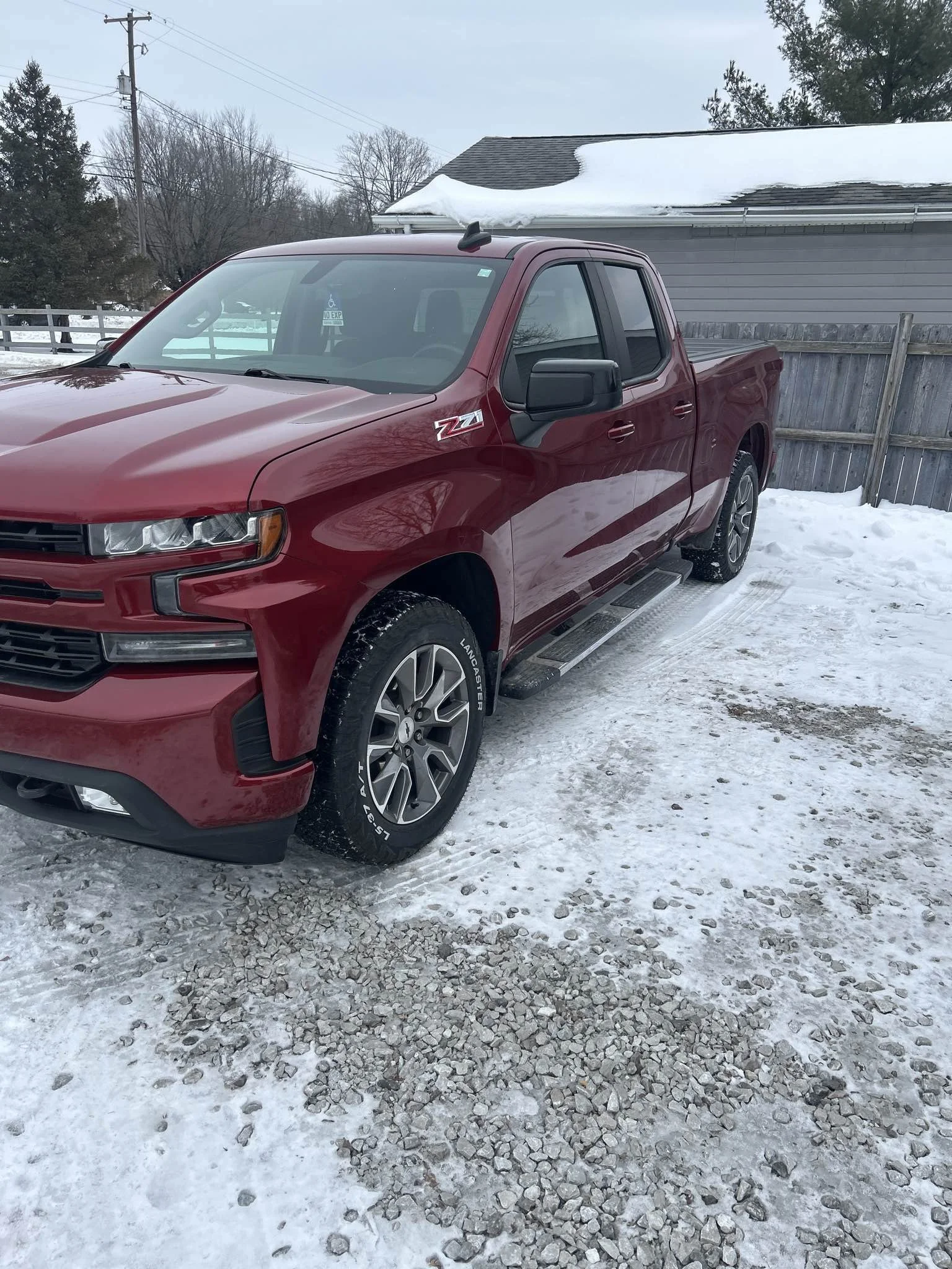Red pickup truck parked on snow-covered ground next to a wooden fence and house with snow on the roof, trees and utility poles in the background.