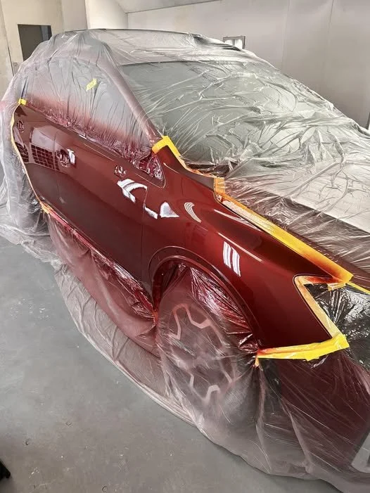 A red car being painted in a workshop, covered with plastic sheeting and masking tape.