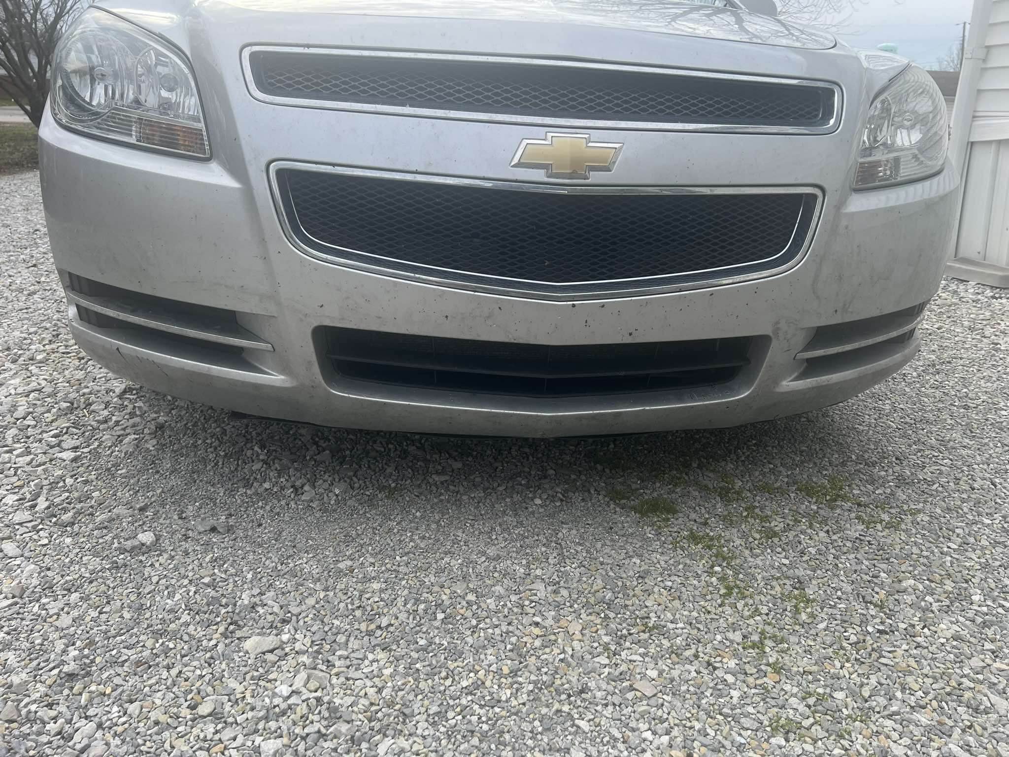 Front view of a silver Chevrolet sedan parked on a gravel surface, showing the grille, headlights, and bumper.