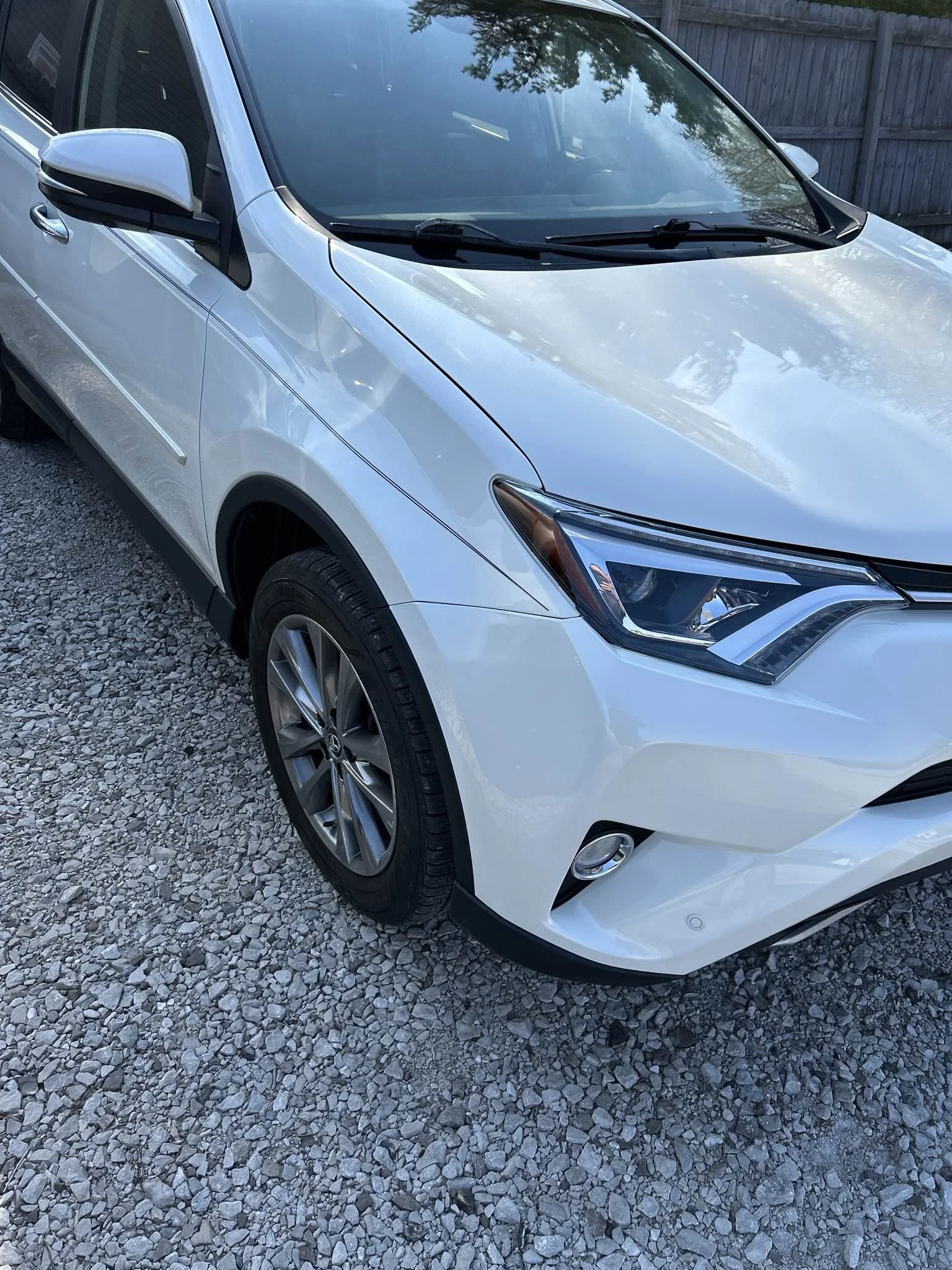 Front left side of a white Honda CR-V SUV parked on gravel with a wooden fence and trees in the background.