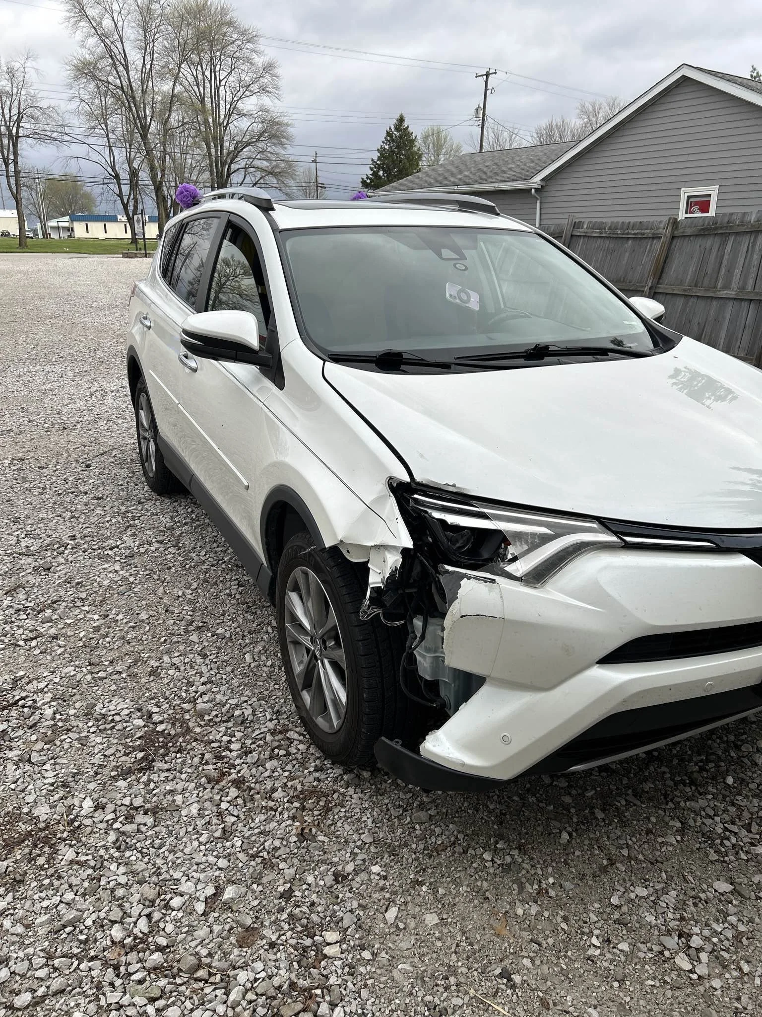 White SUV with front-left damage and missing headlight parked on gravel driveway.