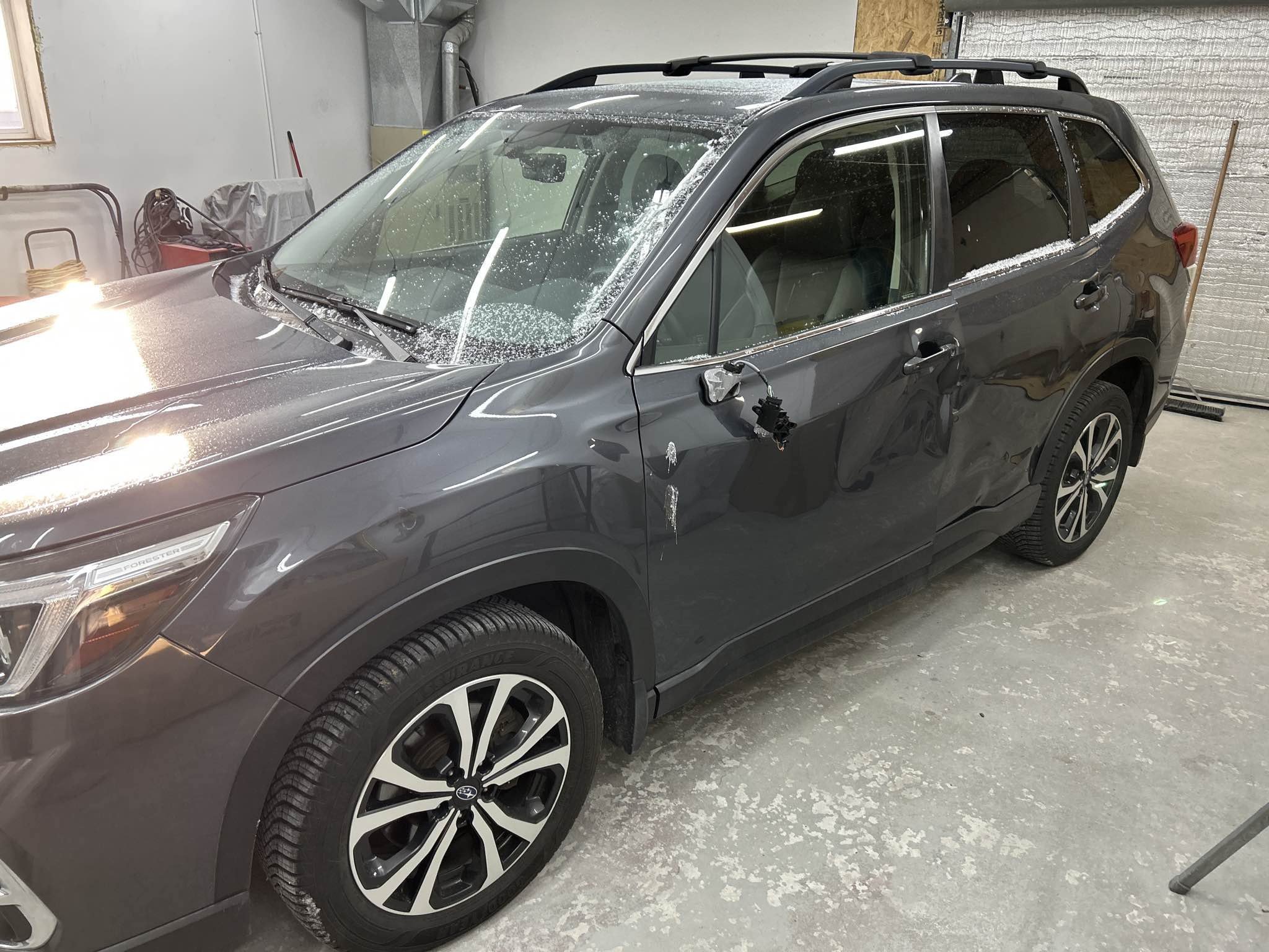 A dark gray SUV with a cracked driver side mirror and damaged driver side door, parked inside a garage. The SUV appears to have a dent on the driver side door and some ice or frost on the windows and roof.