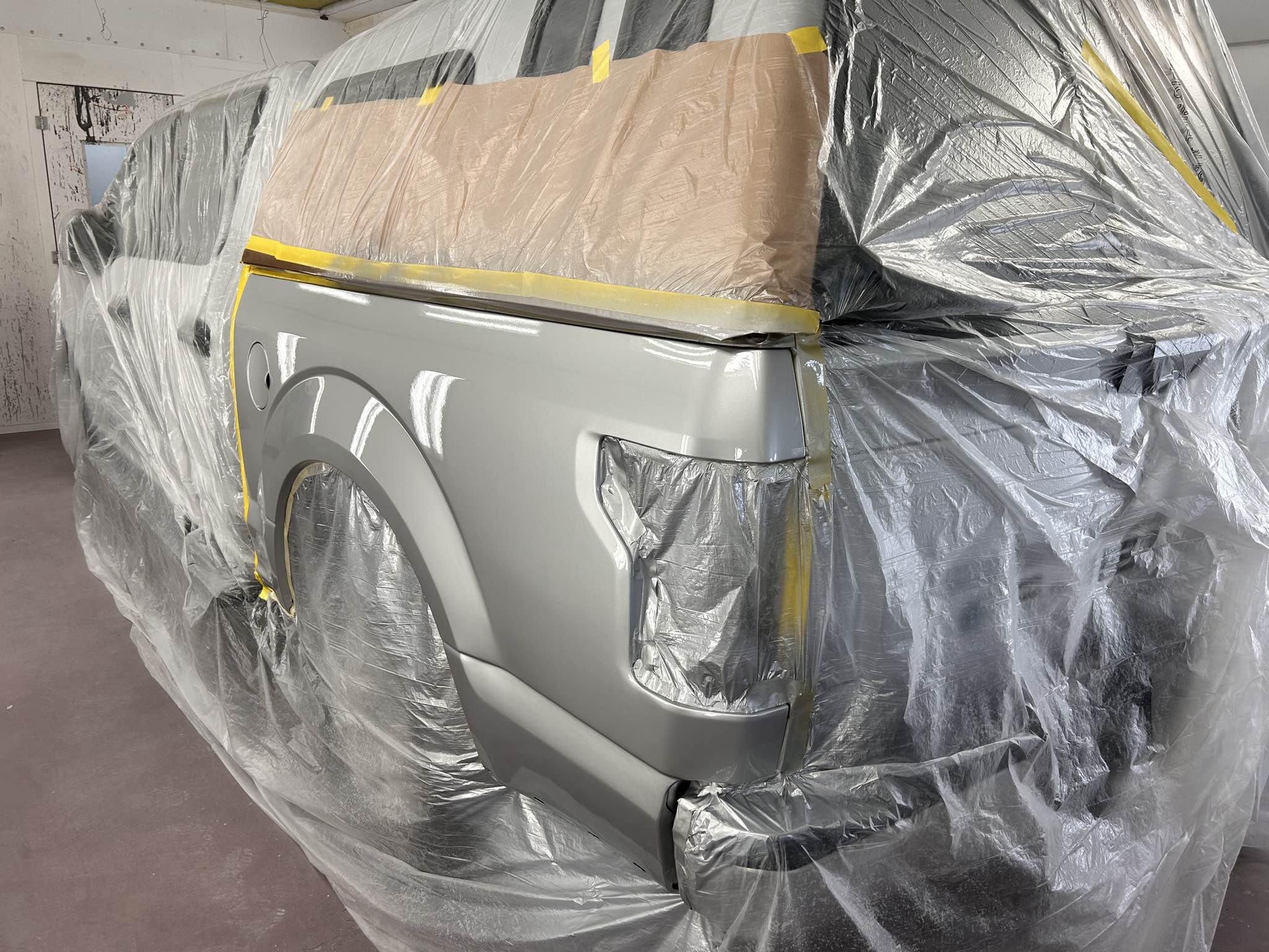 Car in a paint booth covered with plastic and masking tape, being prepared for painting.