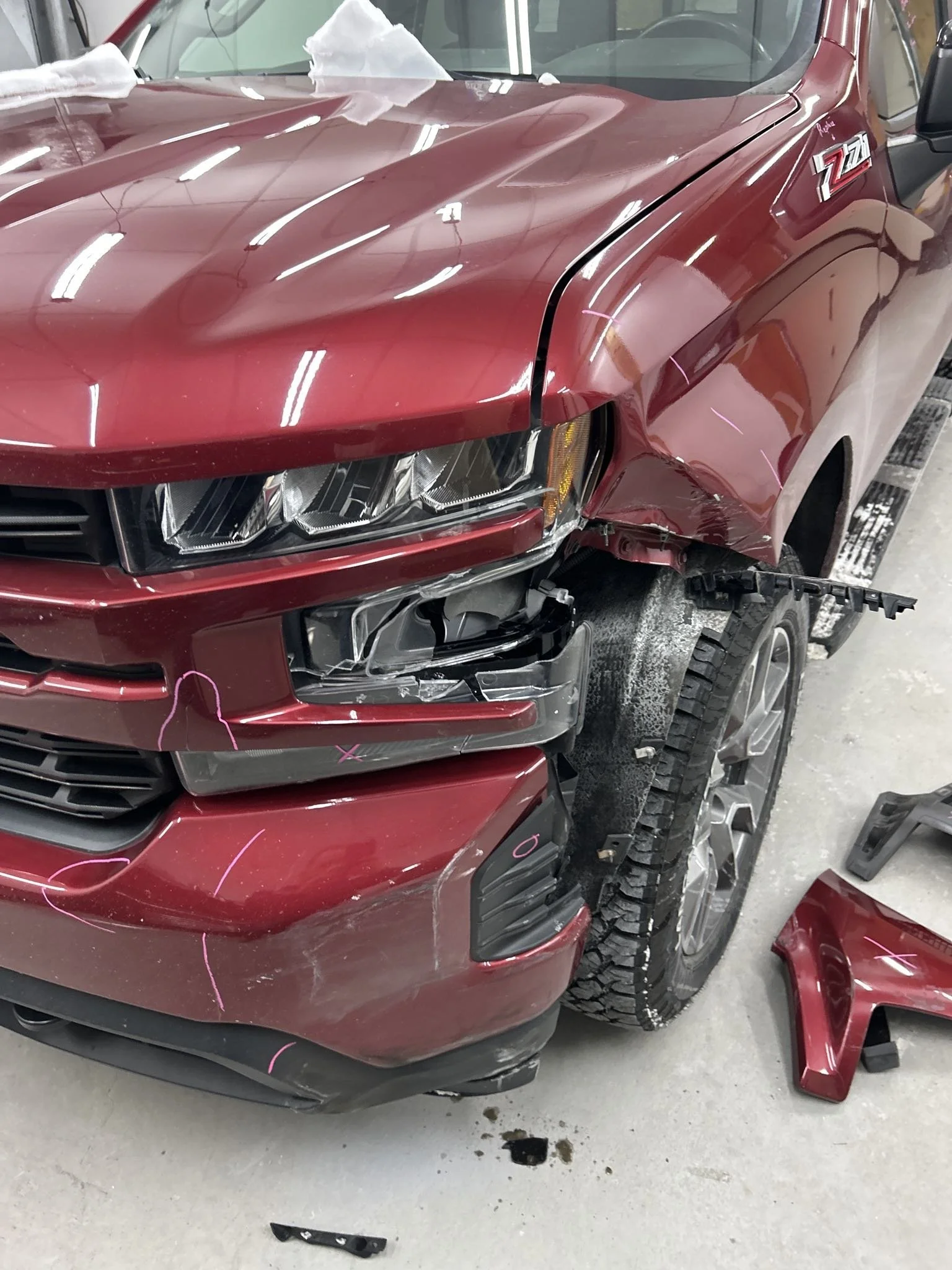 Red pickup truck with front-end damage and parts lying on the floor inside a garage.