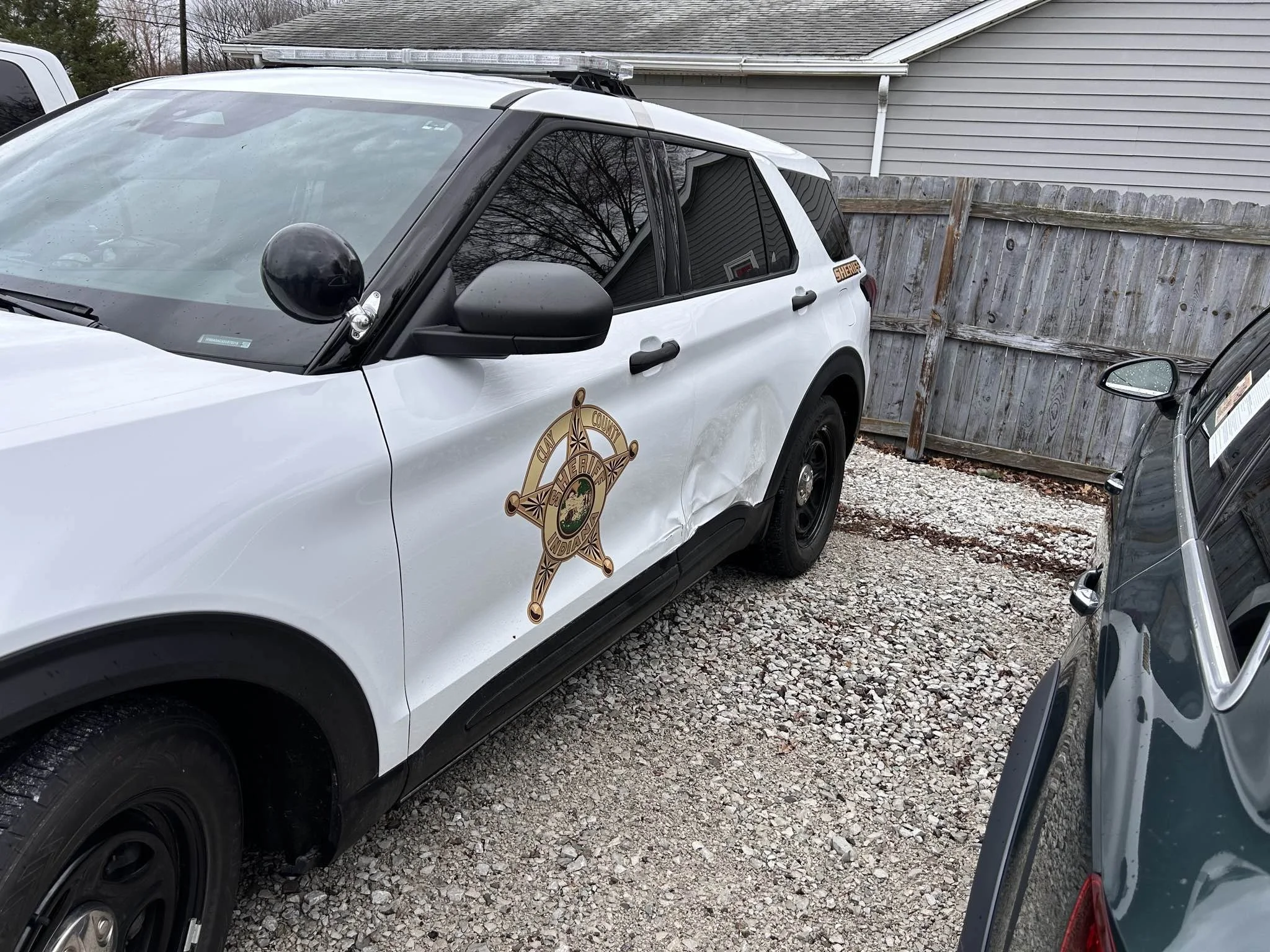 A white sheriff vehicle with a black side mirror and a sheriff badge emblem on the door, parked on a gravel driveway next to a gray house and wooden fence.