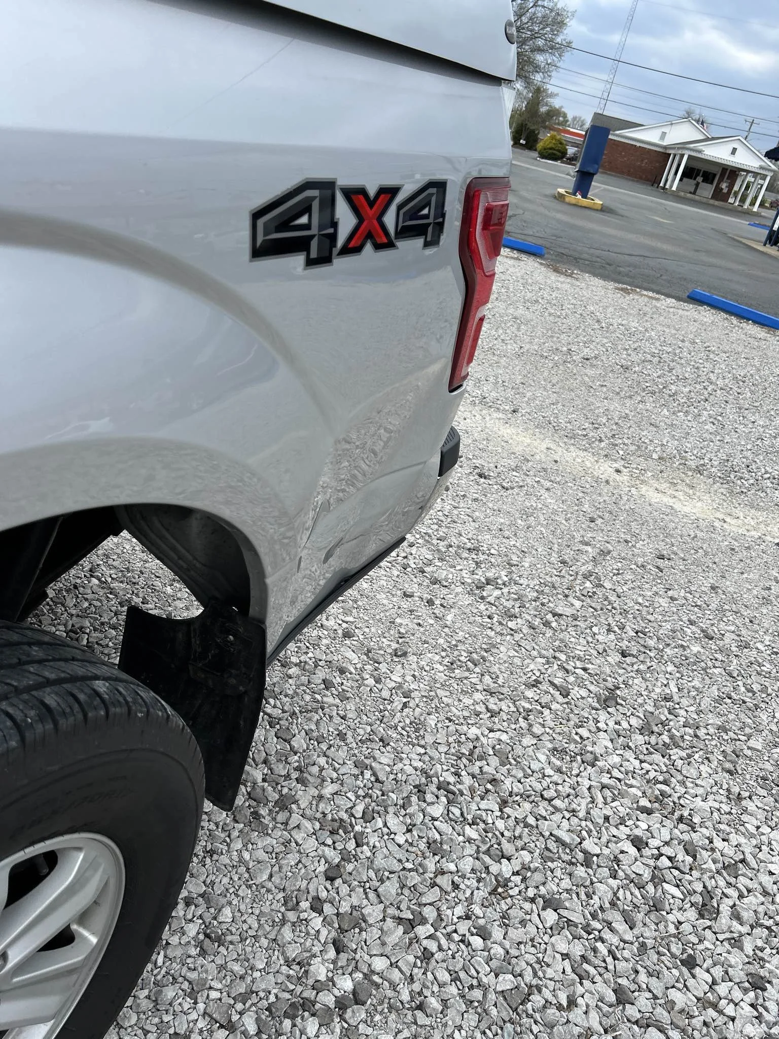 Close-up of the rear side of a white pickup truck with a red and black 4x4 emblem, parked on gravel near a parking lot with a small commercial building in the background.