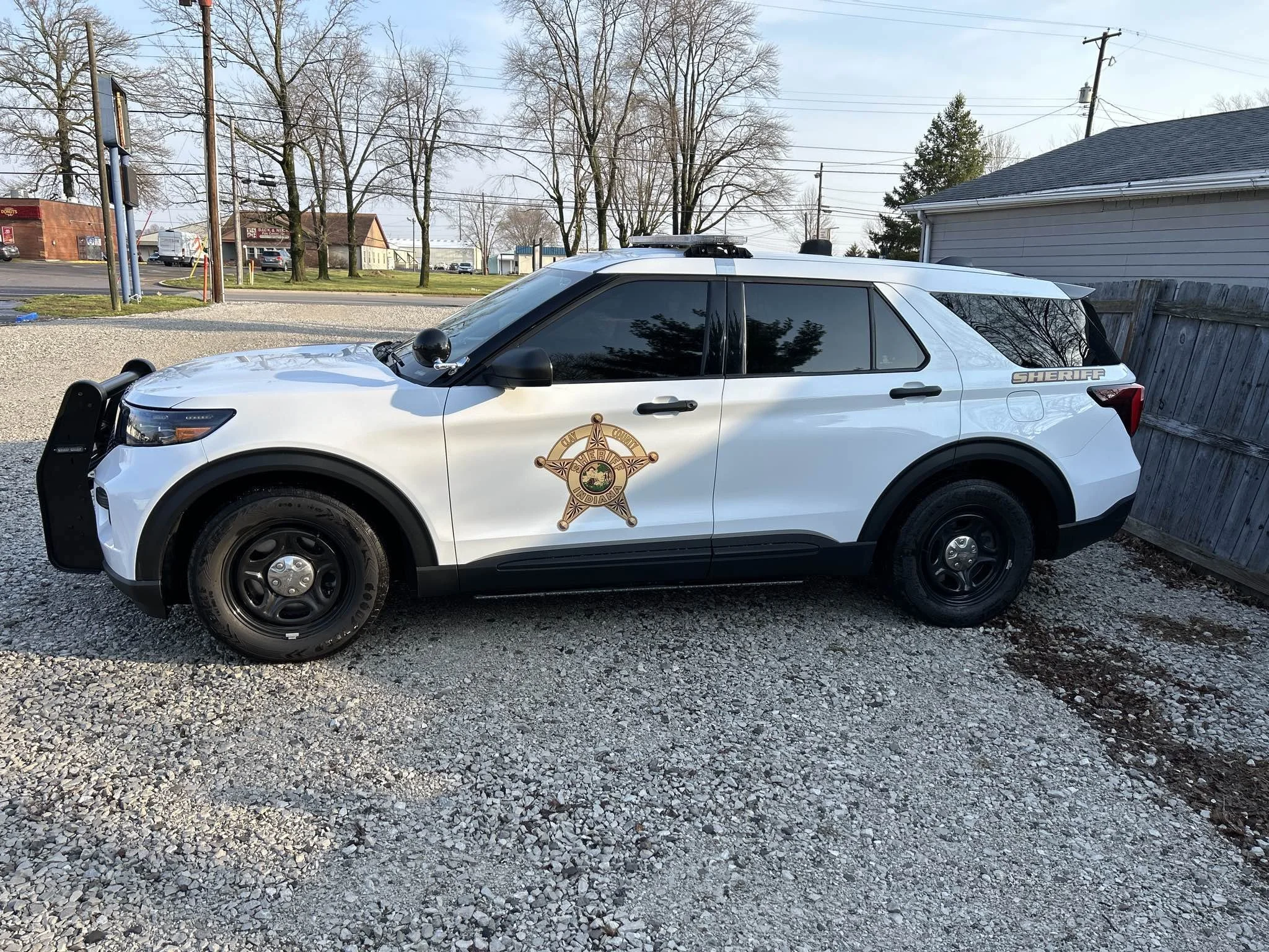 White sheriff SUV parked on gravel lot next to a wooden fence with a sheriff badge emblem on the door.