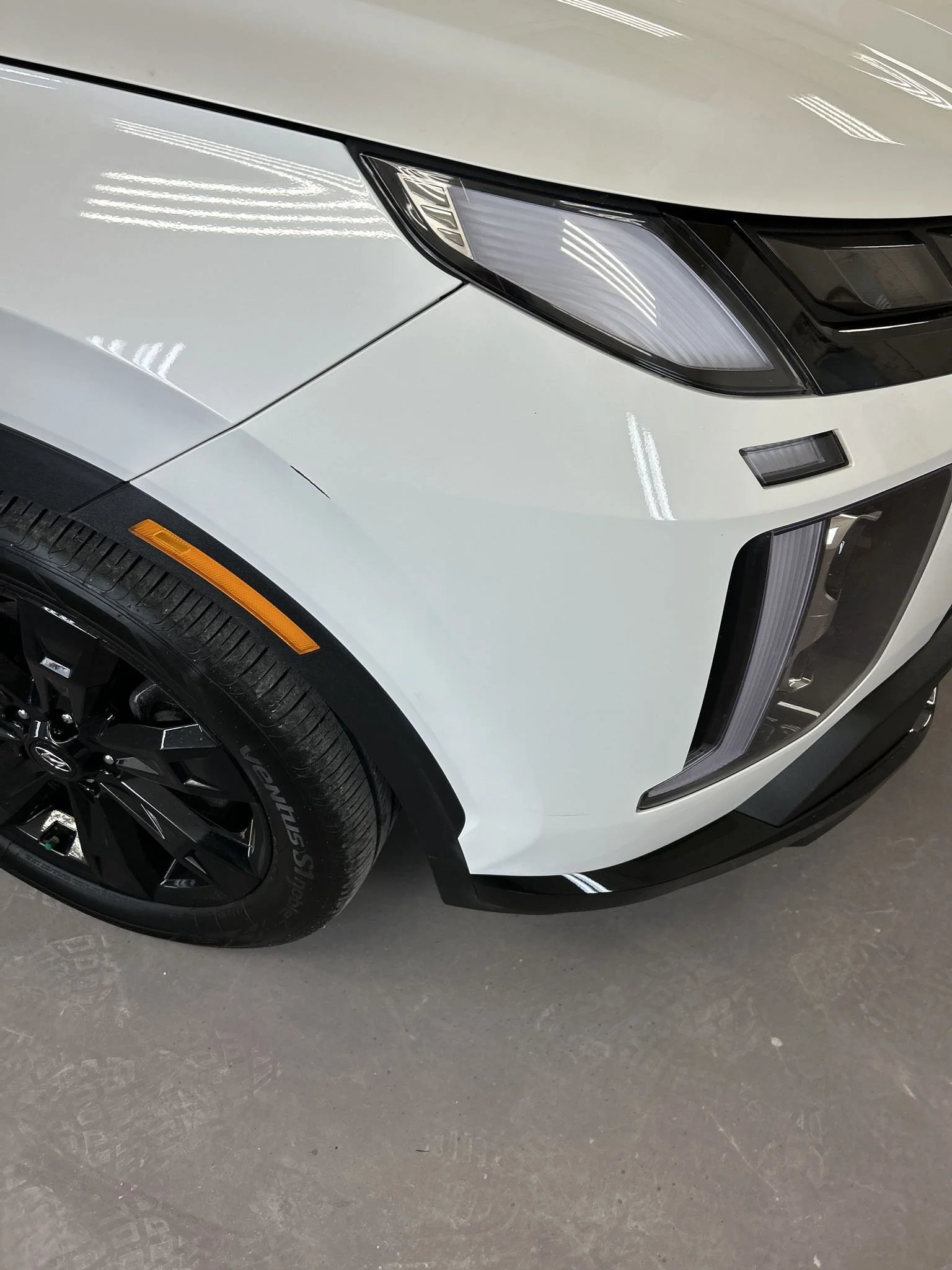 Close-up of the front part of a white modern sports car, showing the headlight, front bumper, and front wheel with black alloy rim and Vision tires, inside a showroom with reflective overhead lights.