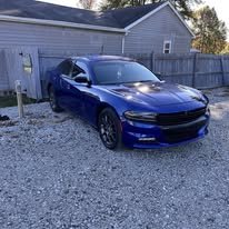 A blue sports car parked on a gravel driveway with a wooden fence and a house in the background.