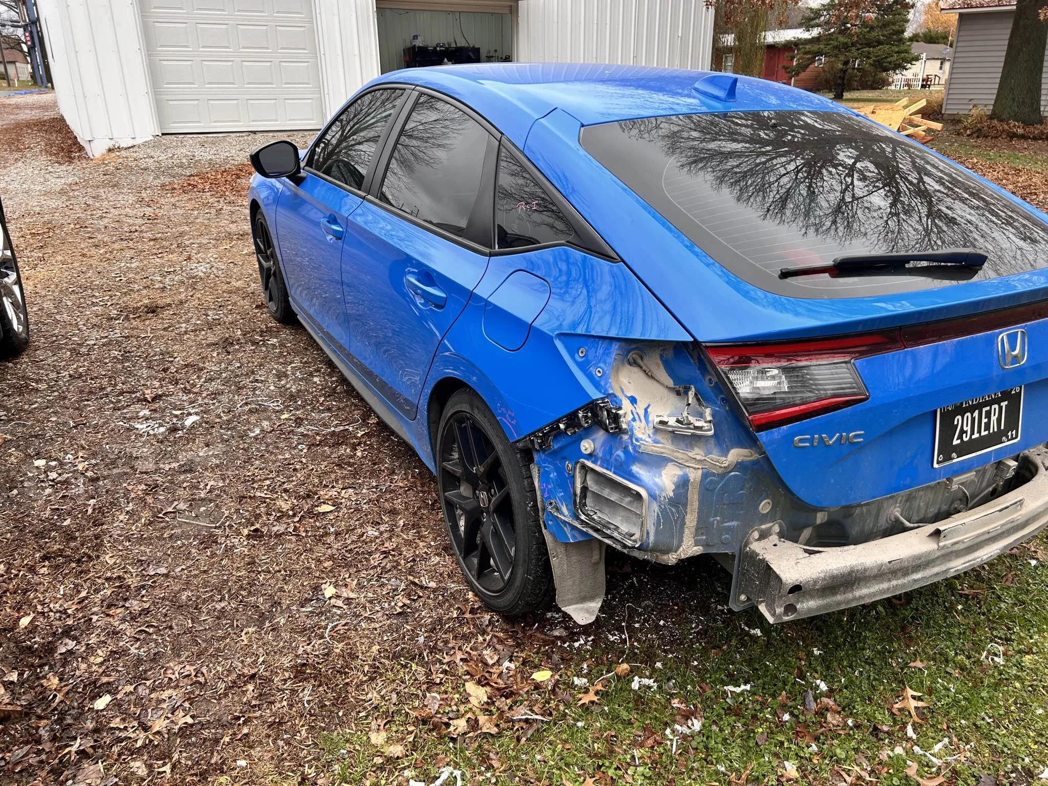 Blue Honda Civic with rear damage parked on a gravel driveway. The rear bumper and tail light area are damaged and parts are missing or disassembled.