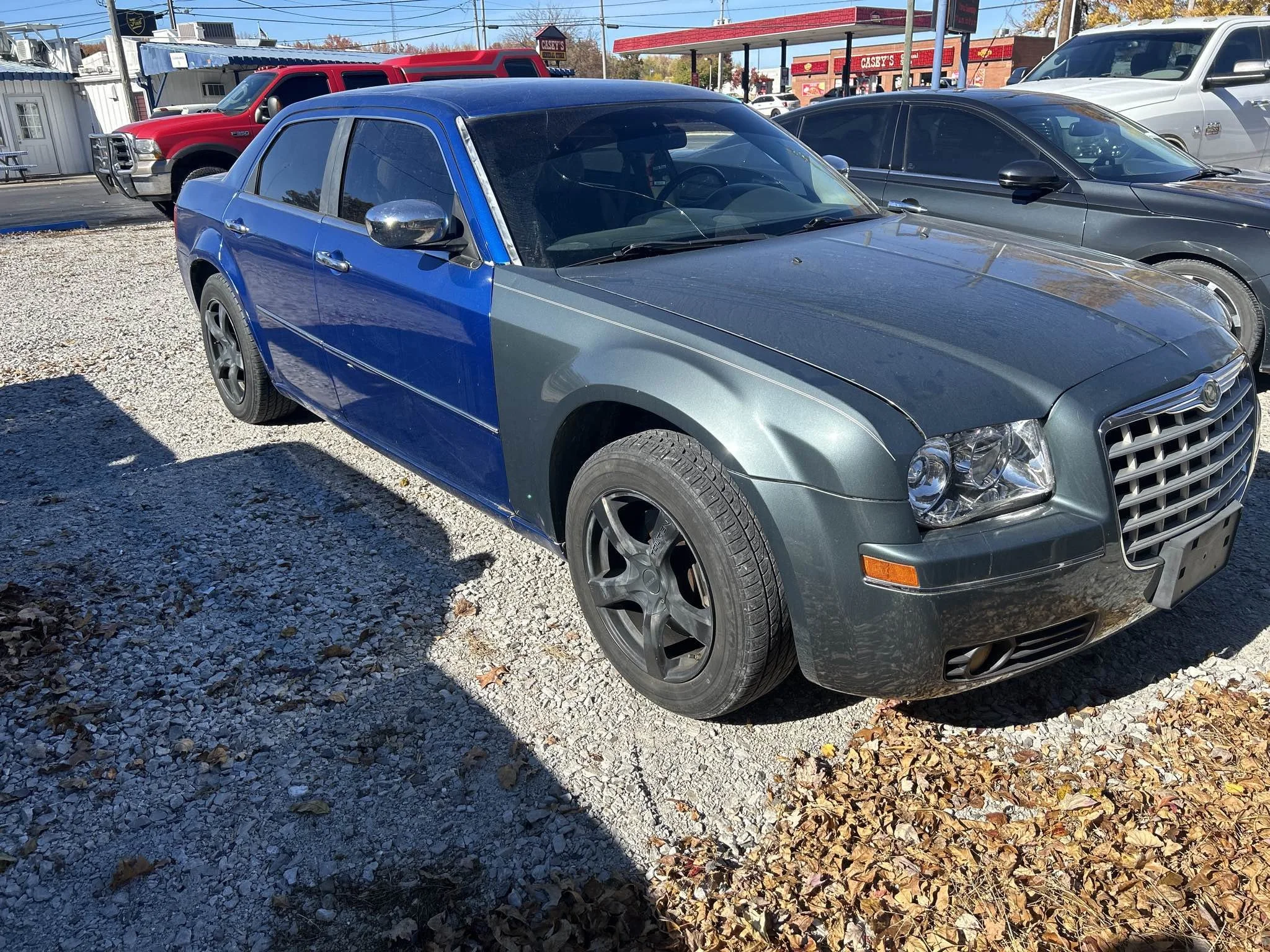 A Dodge Magnum car with a customized two-tone paint job, featuring a dark gray front and a blue rear section, parked on a gravel lot surrounded by other vehicles and buildings.