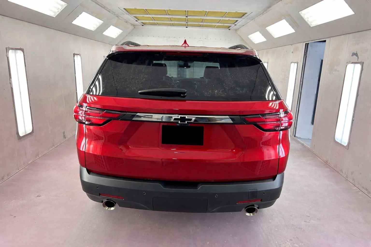 Red Chevrolet SUV inside a spray booth, viewed from the rear, with bright overhead lighting and walls lined with vertical lights.