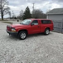 Red SUV parked on gravel driveway next to a wooden fence with trees and cloudy sky in the background.