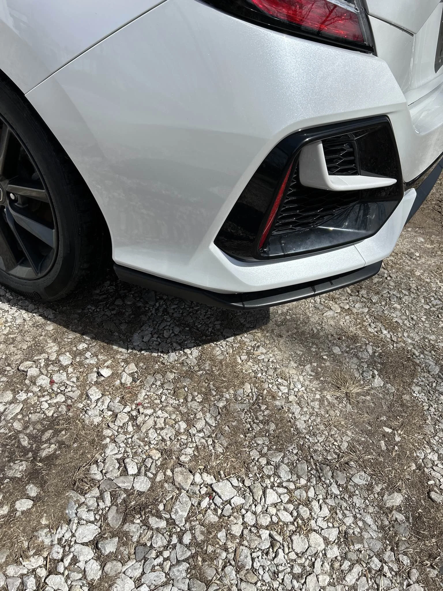 Close-up of the front right corner of a white car, showing the wheel, headlight, and front grille, parked on a gravel surface.