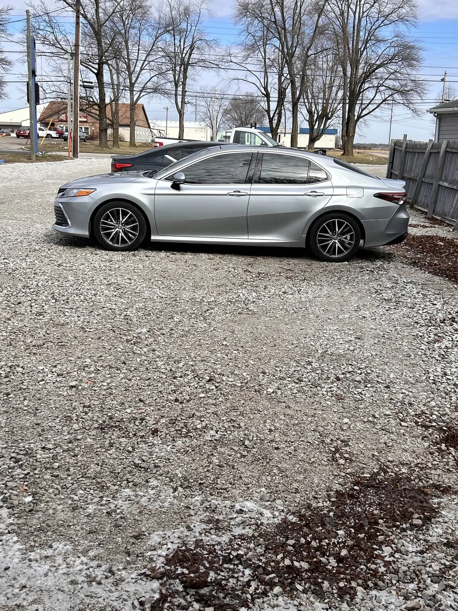 A silver sedan parked on a gravel lot with a black or dark gray car behind it. There are leafless trees, buildings, and power lines in the background.