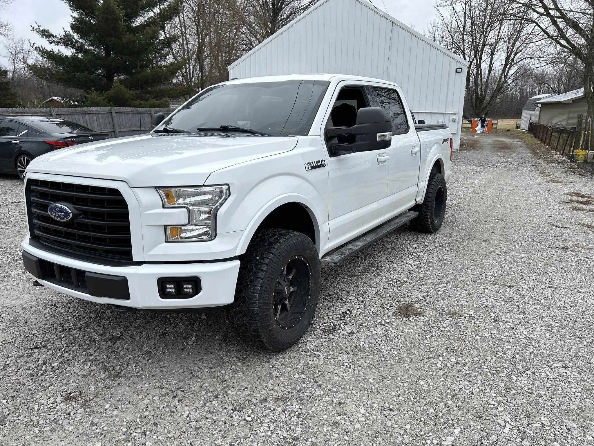 White Ford F-150 pickup truck parked on a gravel driveway with a large white building and trees in the background.
