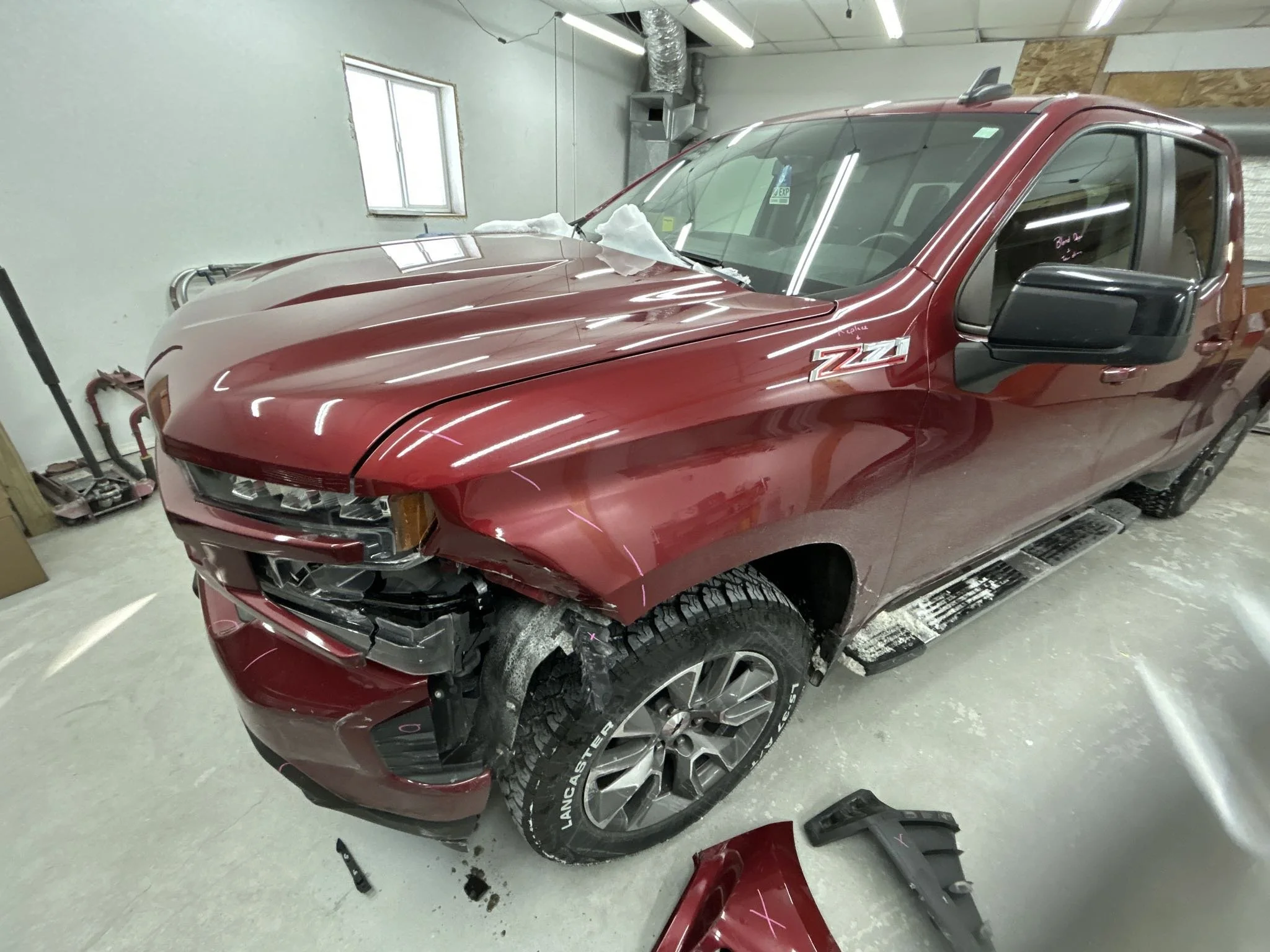 Red pickup truck with front-end damage, missing bumper, and part of the front panel removed, inside a garage.
