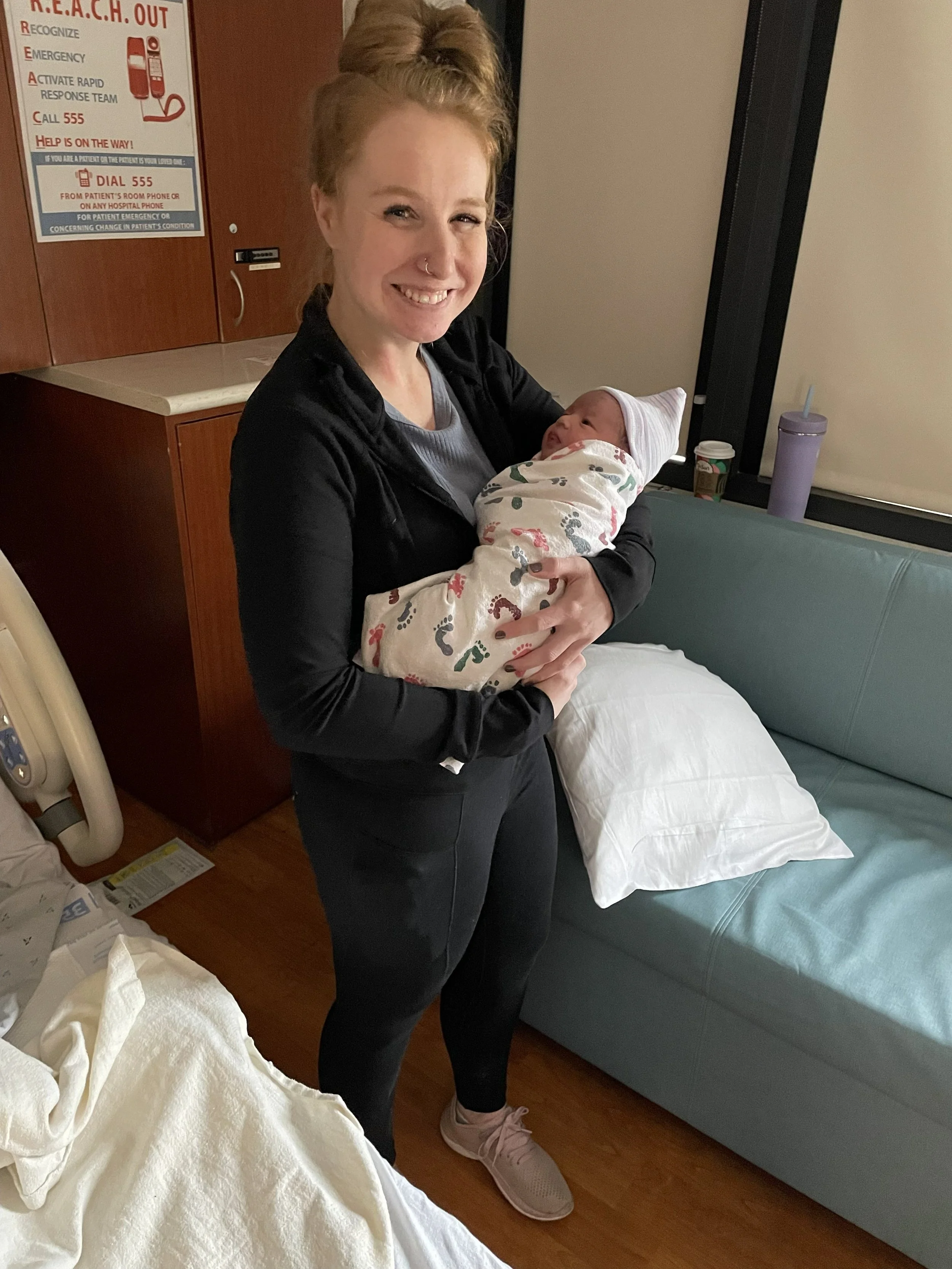A woman holding a newborn baby wrapped in a blanket, smiling in a hospital room.