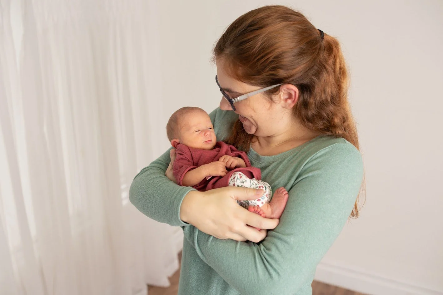 A woman with red hair and glasses holding a newborn baby in a cozy indoor setting.