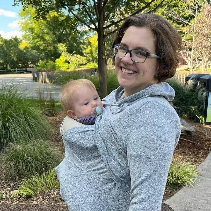 A woman with glasses smiling and holding a baby with a pacifier in a gray hoodie carrier outdoors. Green trees and grass in the background.