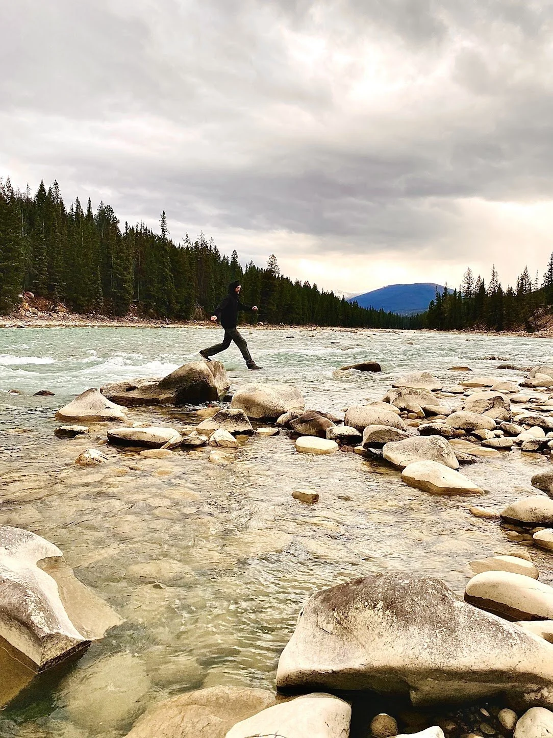 A person in black clothing crossing a rocky river with a forested mountain background under cloudy skies.