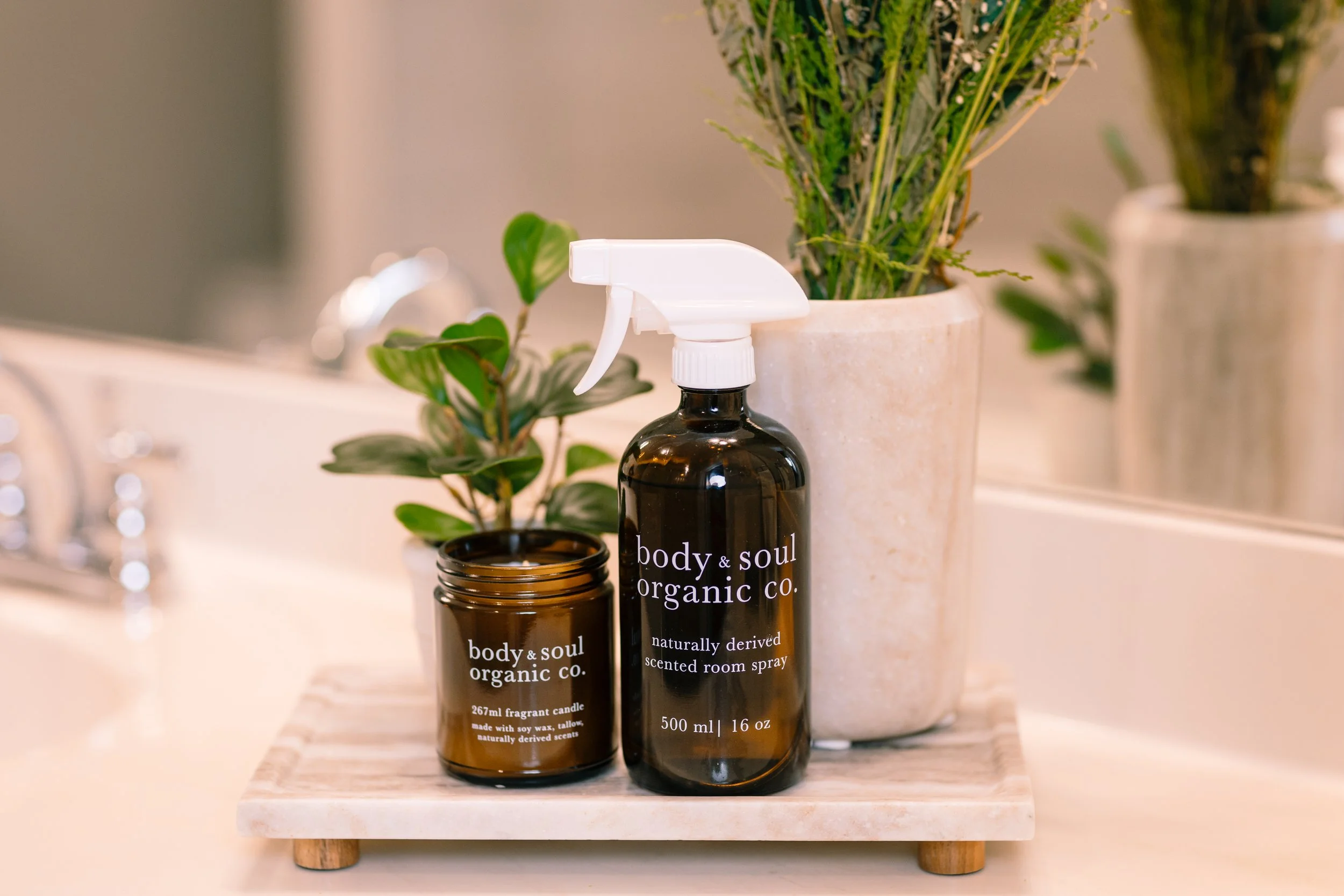 Top view of a bathroom countertop with two dark amber glass bottles labeled 'body & soul organic co.' containing scented room spray and candle, a small potted green plant, and a larger potted green plant in the background.