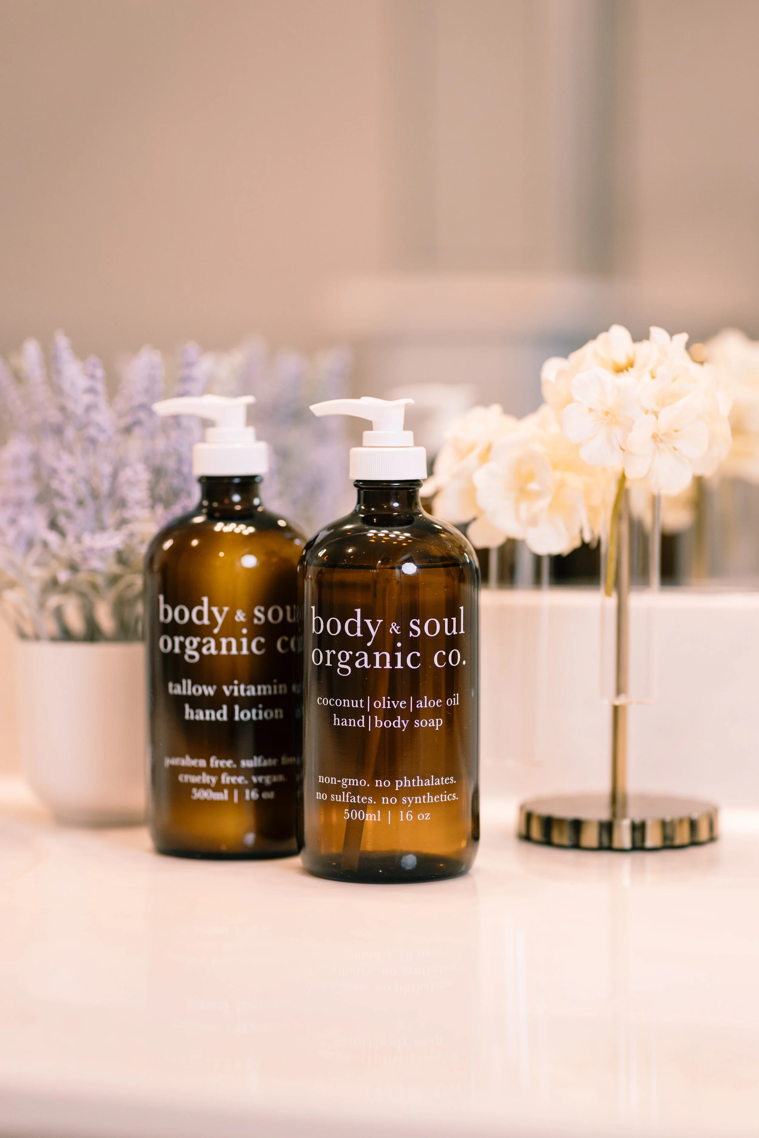 Two amber bottles with white pump caps labeled 'body & soul organic co.' standing on a white surface, with white flowers in a glass vase and lavender in the background.