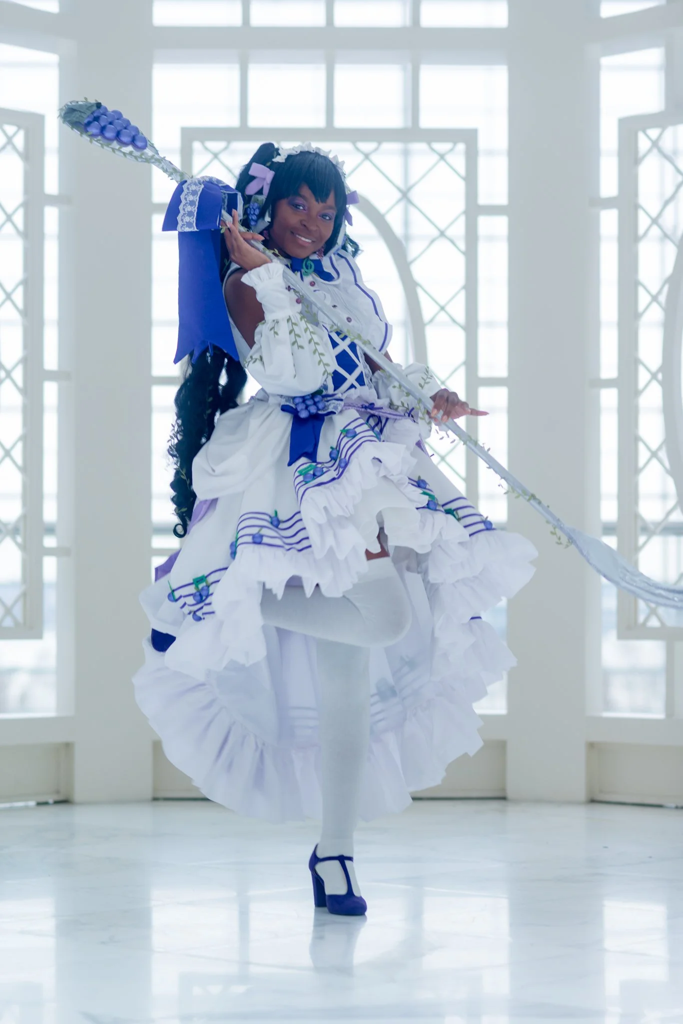 Woman in a blue and white dress with elaborate ruffles, holding a staff with grapes on top, in a bright, airy indoor space with large windows.