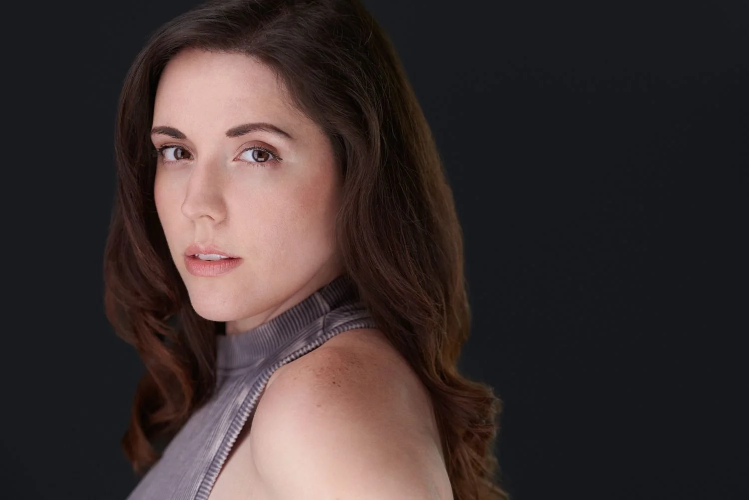 A woman with long brown hair and fair skin, looking over her shoulder at the camera, against a dark background.