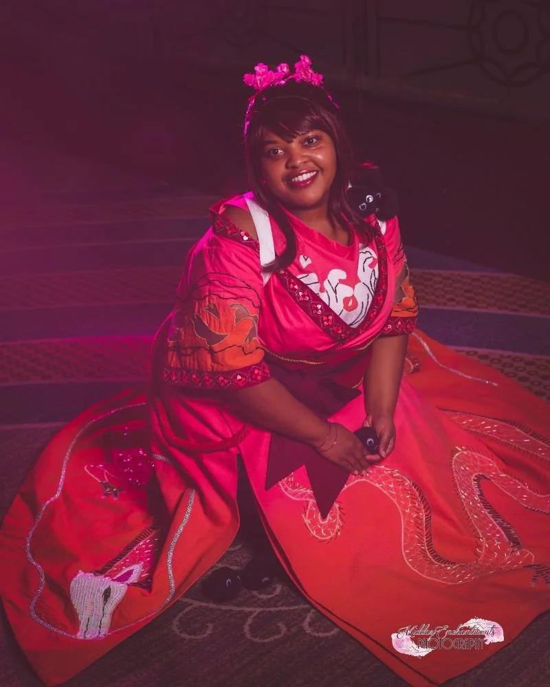 A woman dressed in a vibrant pink and red traditional dress with dragon embroidery, sitting on the floor with a smile, and wearing a pink floral headpiece.