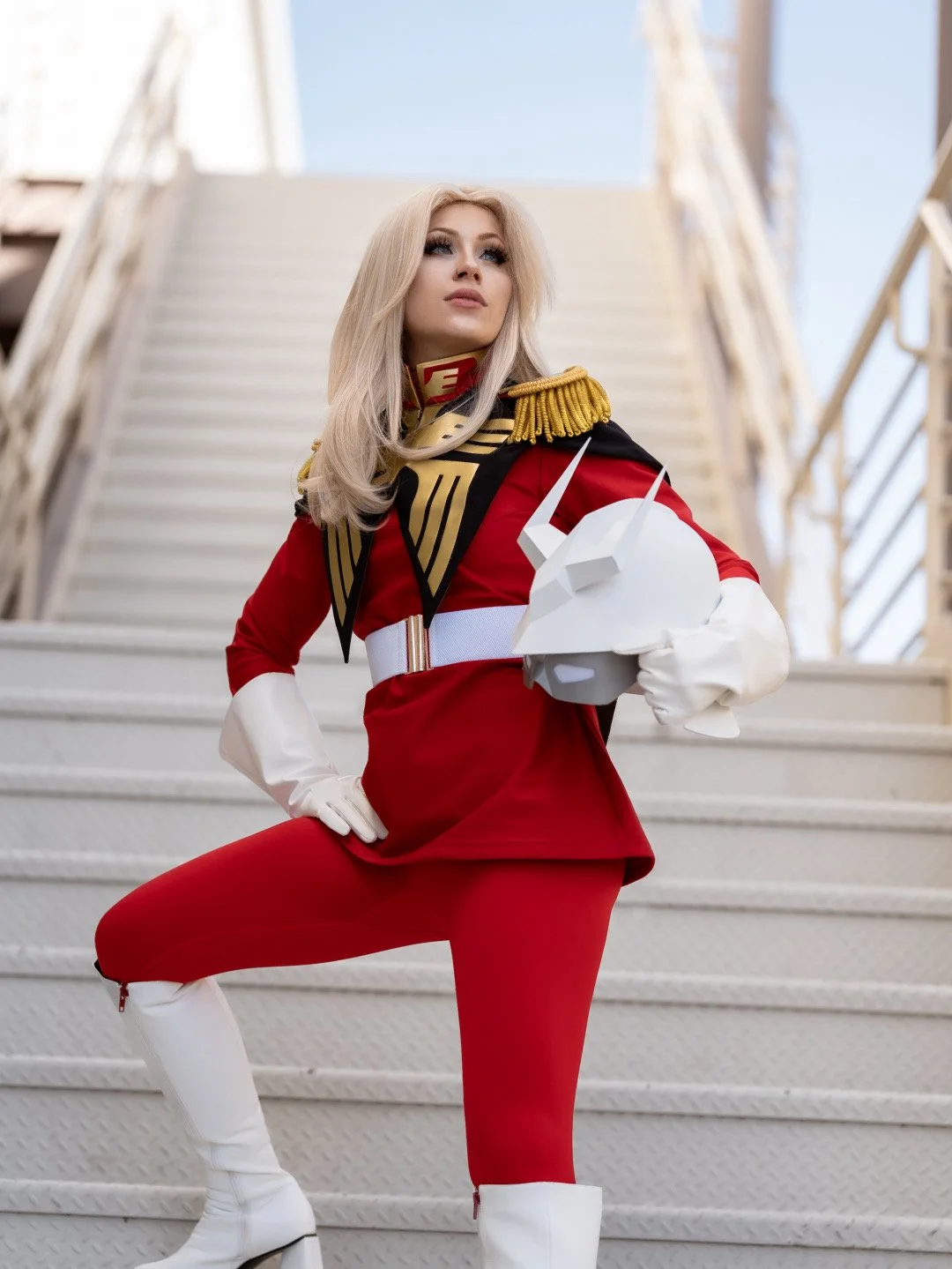 A woman in a detailed red and white costume holding a white helmet with a unicorn horn, standing on stairs outdoors.