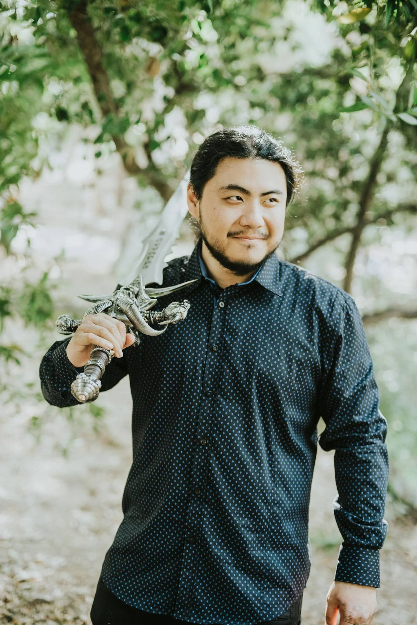 A man with dark hair and a beard, smiling slightly, holding a silver sword with a decorative hilt over his shoulder while standing outdoors surrounded by green trees.