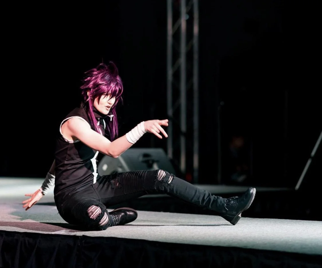 Performer with purple hair sitting on stage in a dynamic pose during a performance.