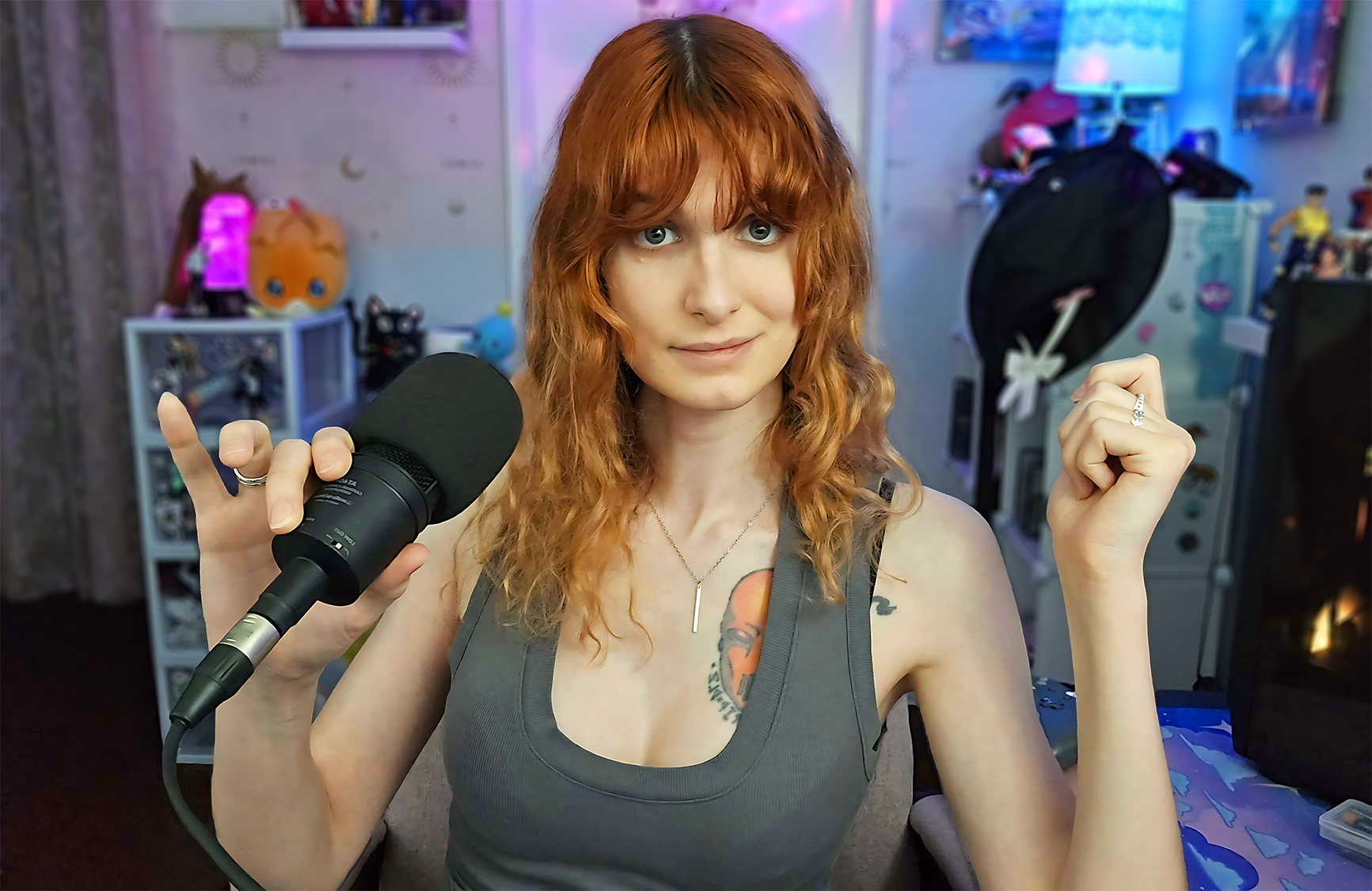 A woman with red curly hair, light blue eyes, wearing a gray tank top and jewelry, holding a microphone in her right hand and raising her left fist, sitting in a colorful, decorated room with toys and vibrant lighting.